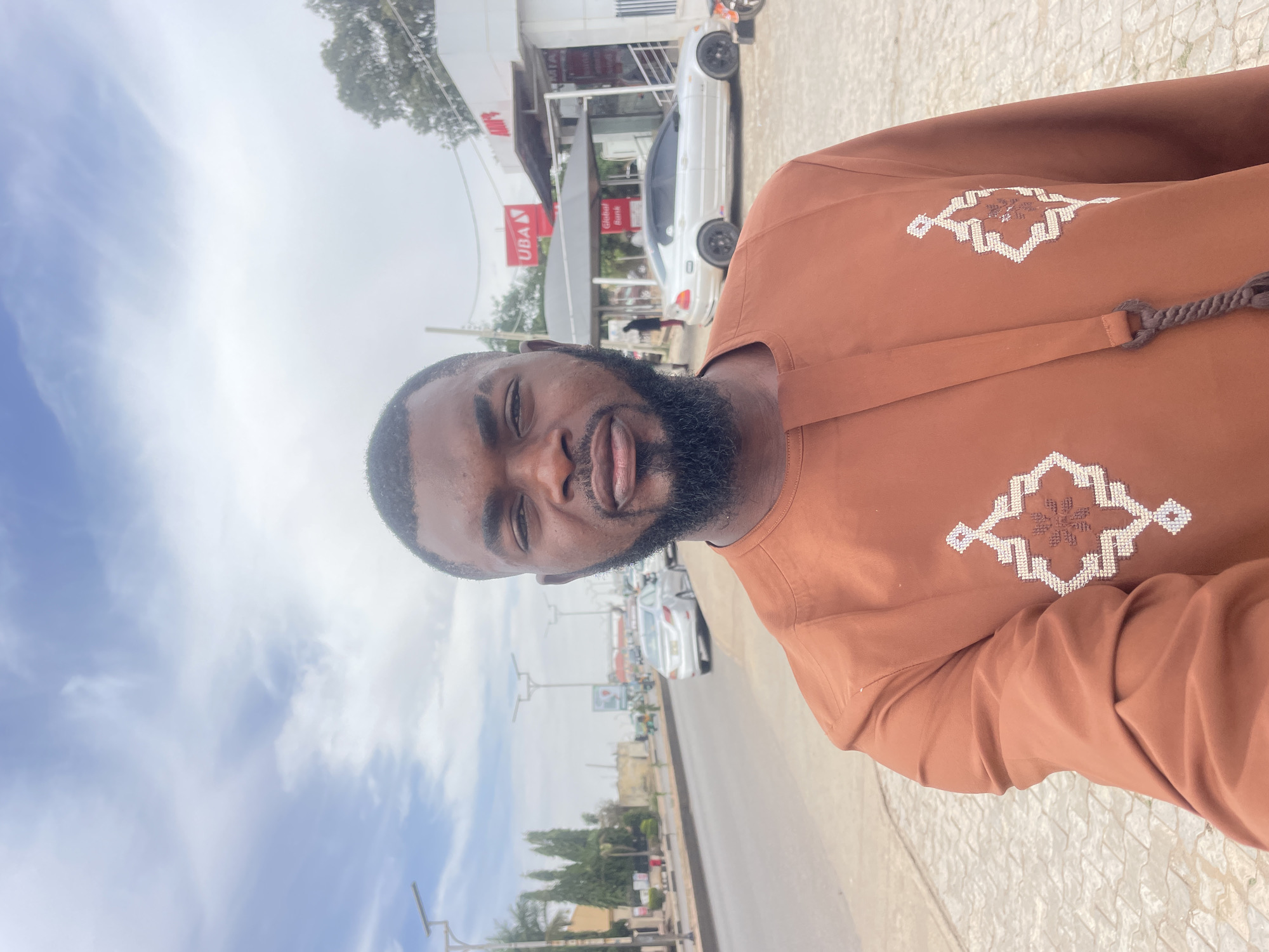 A bearded man wearing a brown tunic with white embroidered geometric designs stands on a sidewalk. Behind him is a street with parked cars and cars moving in the distance. A building with a UBA sign and a "Global Bank" sign is also visible. The sky is cloudy and the photo is taken outdoors. The scene appears to be in an urban area in Jos, Nigeria.