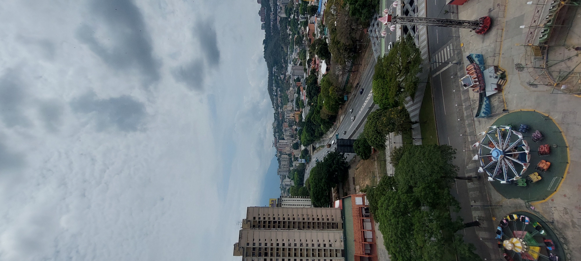 The image is an aerial view of Caracas, Venezuela, taken on an overcast day. The city sprawls along a valley, ascending up a hillside. A major road runs through the cityscape, lined with trees. A building with an unfinished facade and a red roof is visible in the foreground. A nearby amusement park features several rides. Traffic is visible on the road.