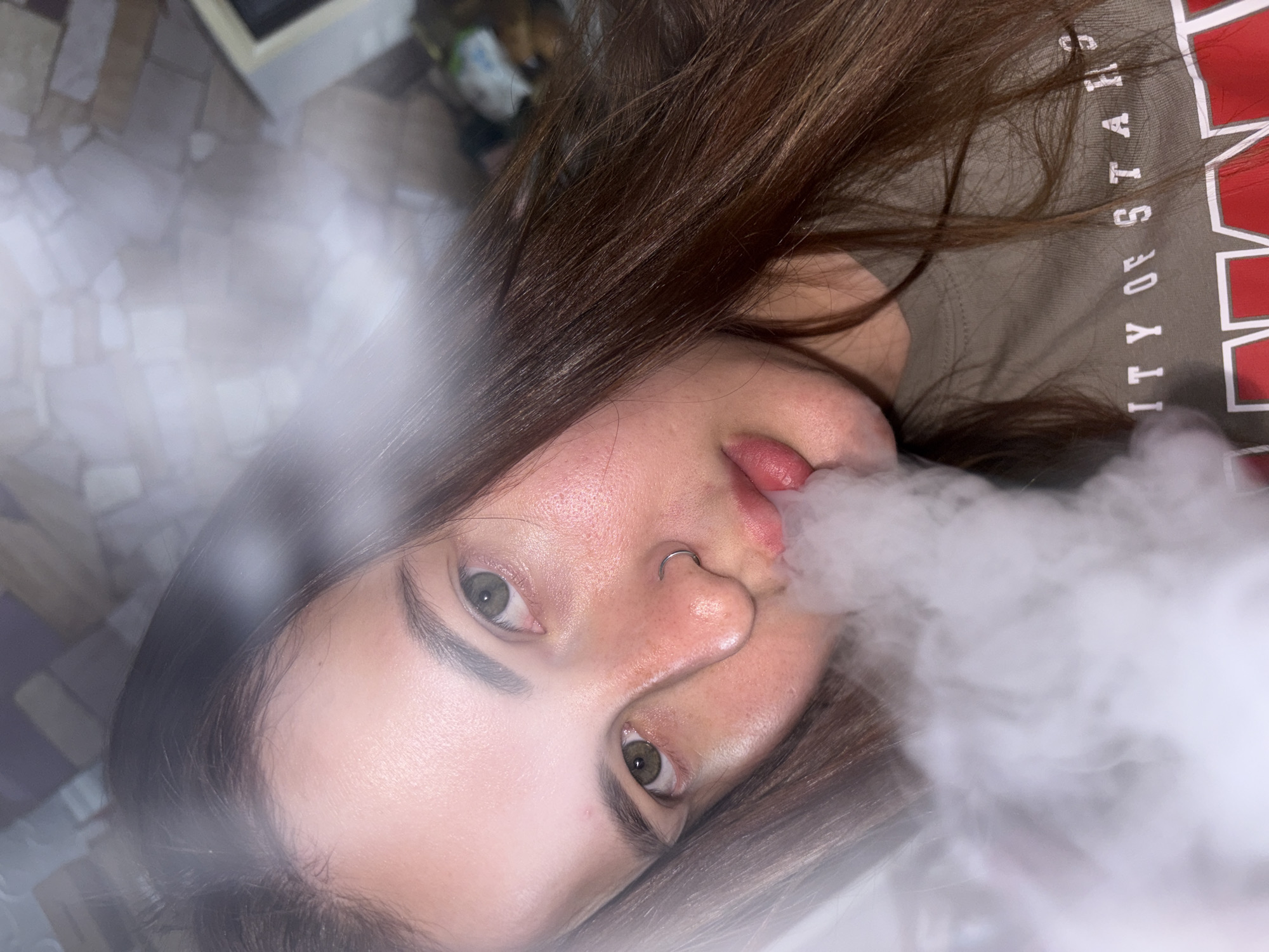 A close-up, angled shot features a person exhaling smoke. The person has brown hair, light skin, and green eyes, with a nose ring visible. They are wearing a brown t-shirt with the text "ITY OF STARS" in white letters and red outlines. The background is blurred and indistinct, showing what appears to be a tiled floor and a corner of a white object. The photograph does not provide enough information to determine the location as Novopokrovskaya, Russia.