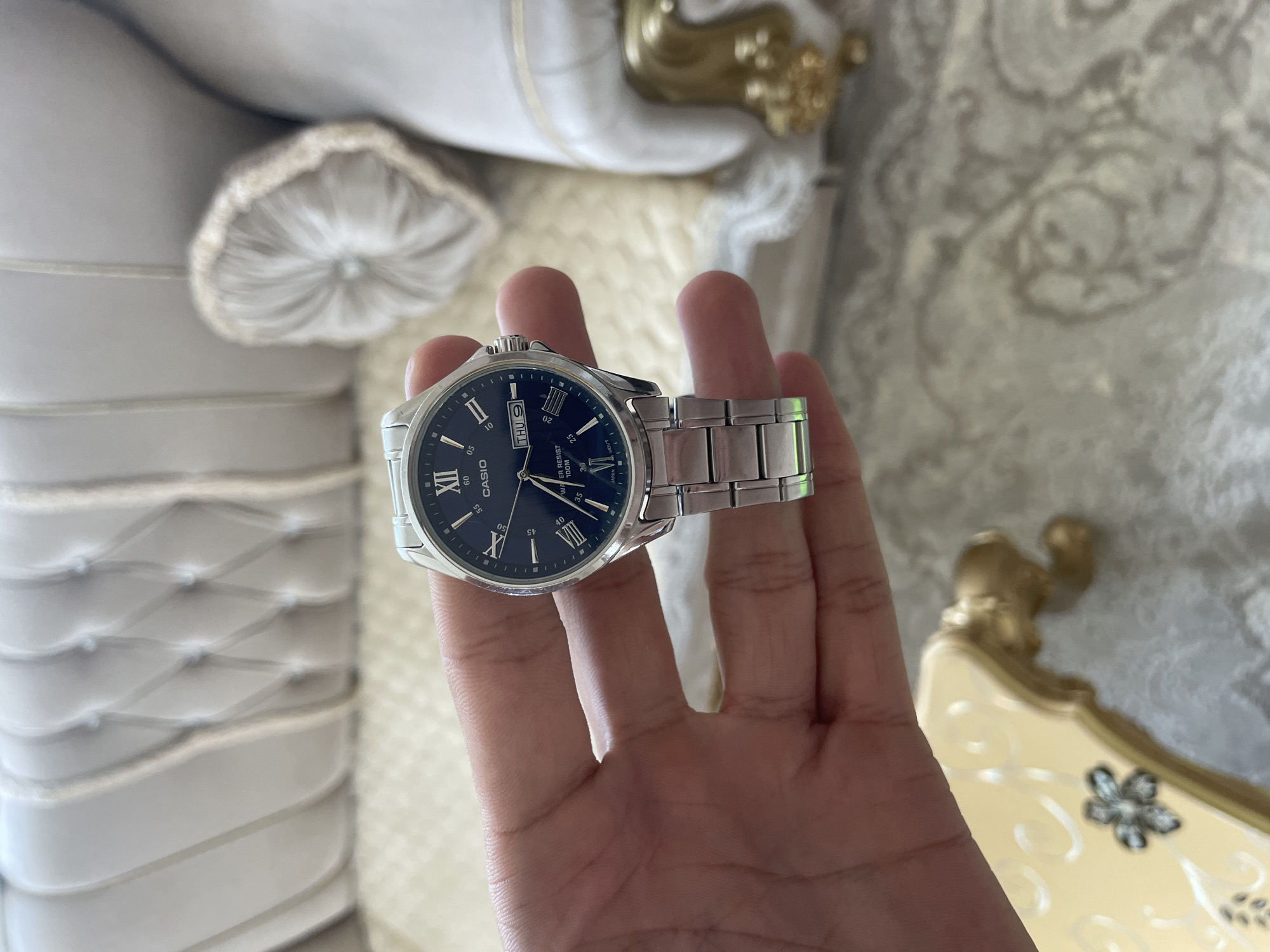 A close-up, eye-level indoor shot captures a hand holding a metallic wristwatch against a softly lit background of ornate, upholstered furniture in Ihtiman, Bulgaria. The central subject is a person's hand, seen from the palm side, presenting a silver-toned wristwatch.
The watch features a dark blue dial with silver Roman numeral hour markers at XII, X, and VIII, and dash markers for the minutes. A day and date window is prominently displayed at the 3 o'clock position, showing "THU 9". The minute hand points just past the "05" mark (or 1 o'clock position in a standard clock face), and the hour hand is positioned between X and XI, indicating approximately 10:07 or 10:08. Visible text on the watch face includes "CASIO" at the 9 o'clock position, "WATER RESIST 100M" below the day/date window, and "JAPAN MOV'T" near the 6 o'clock position. The watch has a matching silver-tone metal link bracelet.
The background is softly out of focus, revealing opulent indoor furniture. To the left, there's a large piece of furniture, possibly a bed headboard or a sofa, with white or light-colored tufted upholstery. Gold, ornate accents are visible on the furniture to the right and top, suggesting a classical or baroque style. A light-colored patterned rug or fabric can be seen beneath the furniture. The overall lighting is bright and even, suggesting daytime.