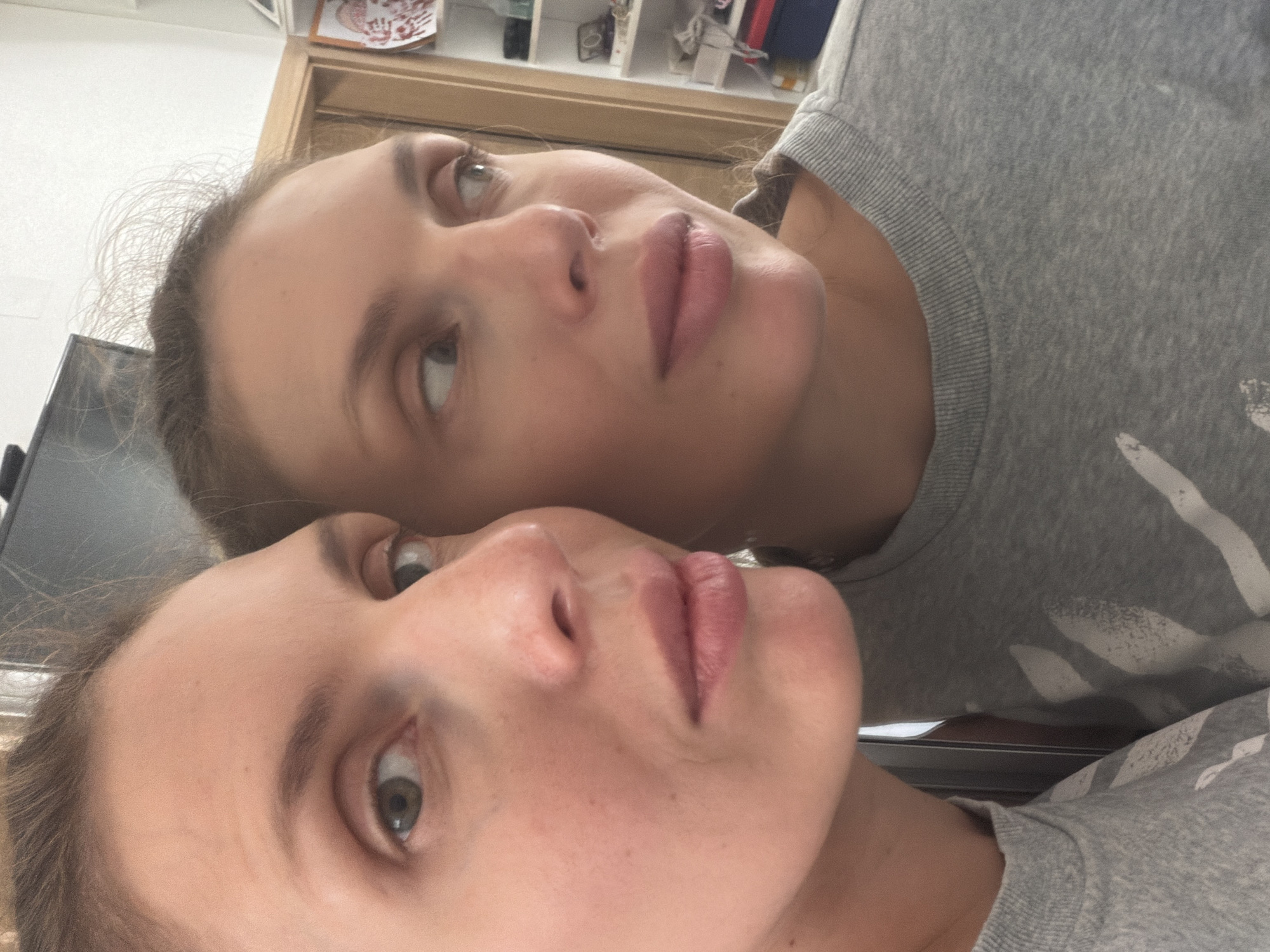 A close-up, reflected profile shot of a fair-skinned woman's face is captured in the foreground. The woman has light brown hair, which appears slightly disheveled, and her gaze is directed to the right, towards a mirror. Her skin shows some subtle texture and pores. She is wearing a gray crew-neck sweatshirt with a white graphic design, portions of which are visible. The background is blurred, but suggests an indoor domestic setting with shelves and what appears to be a doorway. The specific location of Budva, Montenegro, is not discernible from the visual information provided in the image.