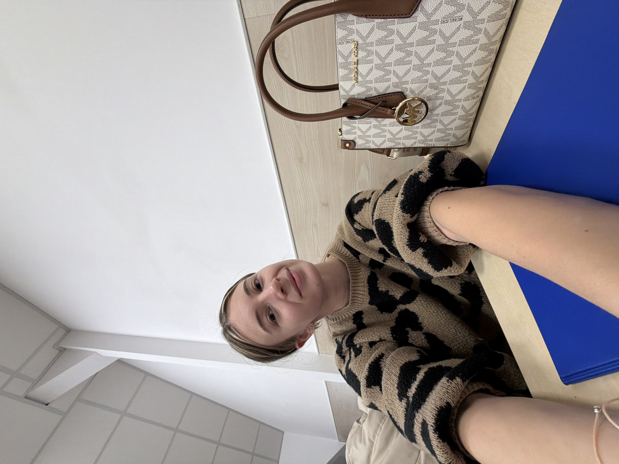 The image shows a person in a black and beige animal print sweater, presumably indoors. A Michael Kors handbag rests on a wooden surface along with a blue folder. The exact location in Plovdiv, Bulgaria is not discernible from the image. There is no interaction evident. The environment appears to be a room, possibly a classroom or office, given the presence of the folder and what looks like a tiled wall in the background.