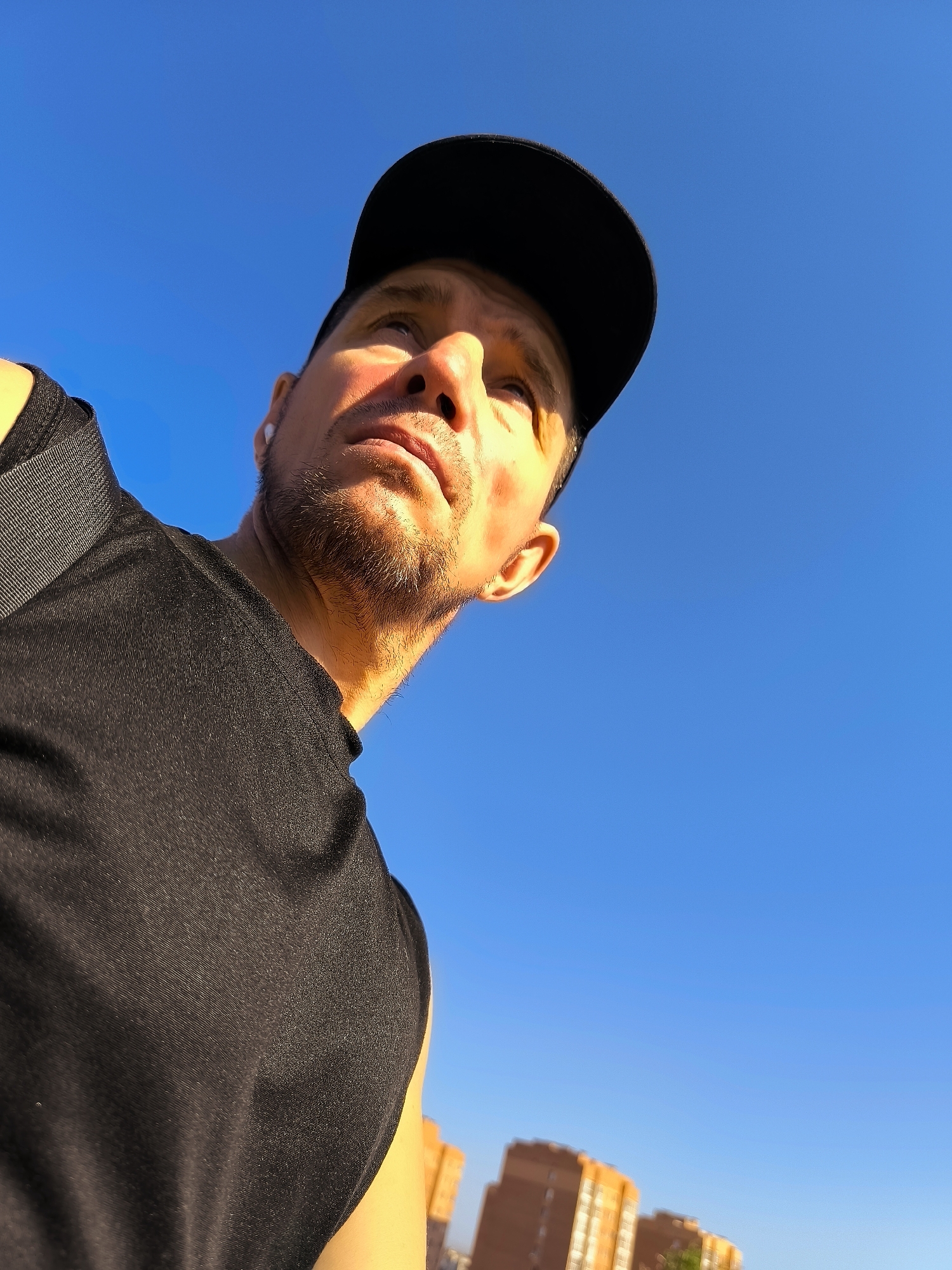 A low-angle, wide shot captures a man wearing a black baseball cap and a black t-shirt, looking upwards against a clear, bright blue sky. Sunlight illuminates his face from the side, highlighting stubble on his chin and the bridge of his nose. He appears to be wearing a white wireless earbud in his visible ear. In the background, two blurry, tall, orange-toned buildings are visible at the bottom of the frame. The scene suggests an outdoor setting, possibly in an urban environment like Qarağandı city administration, Kazakhstan, during daylight.