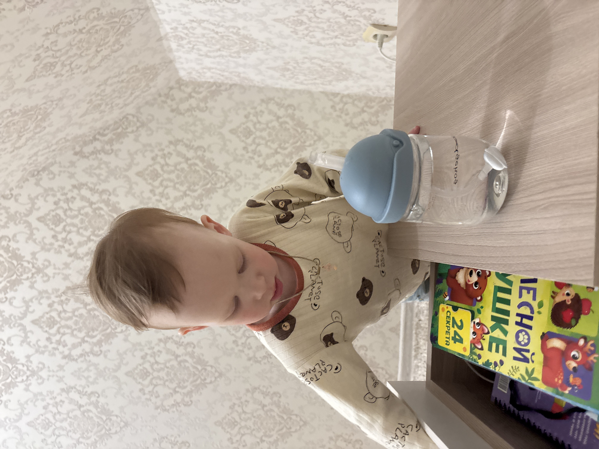 A small child is reaching for a water bottle on a light-colored, wooden surface. The child wears a long-sleeved shirt with bear patterns. A children's book with animal illustrations and text in Russian is visible behind the bottle. The location is likely indoors, given the presence of patterned wallpaper and a wooden surface resembling furniture. The city of Ferma, Russia, cannot be confirmed from the image alone, but it is a possibility.