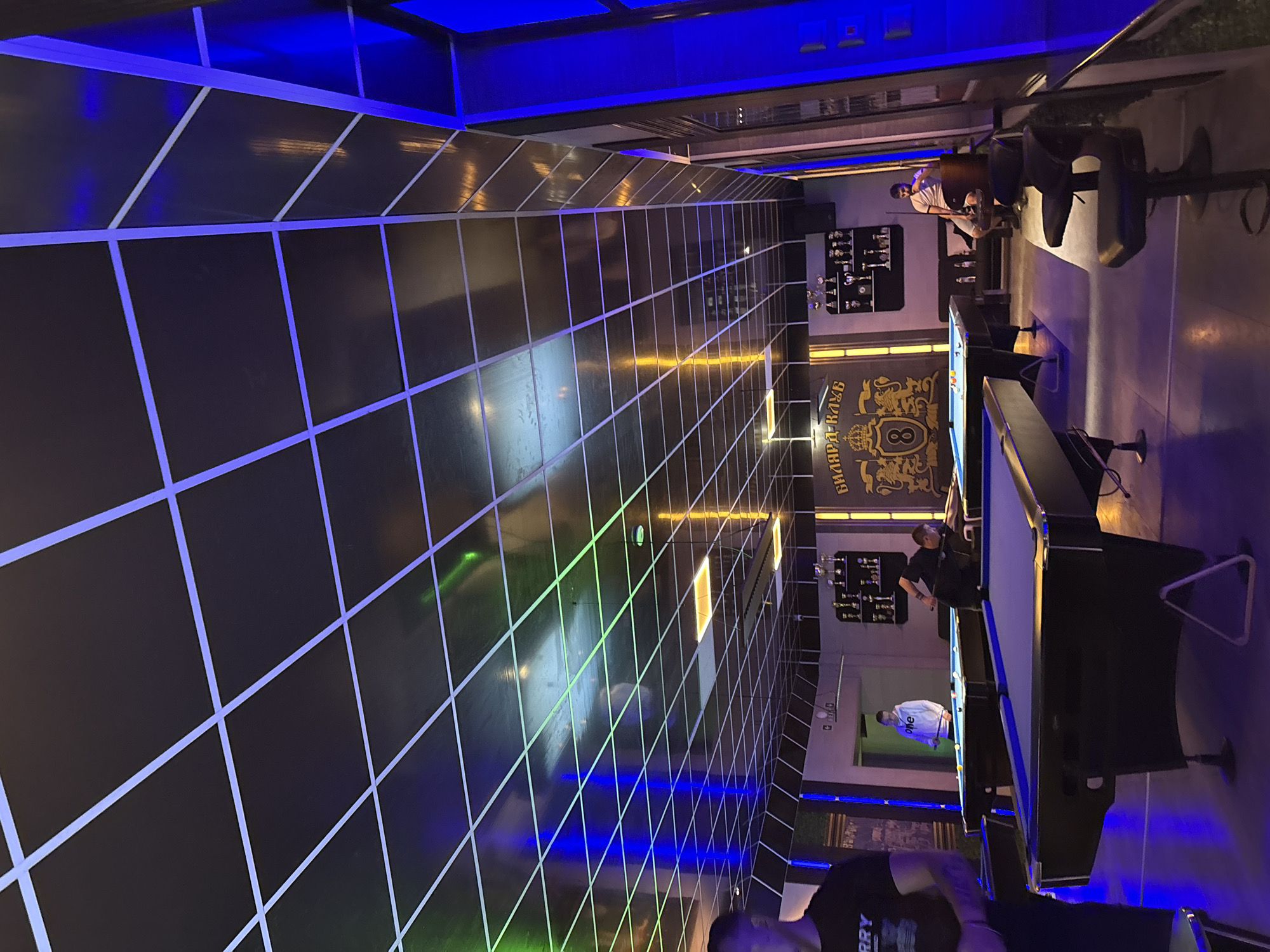 The image depicts the interior of a billiard club in Ruse, Bulgaria. The walls and ceiling are black with a grid pattern formed by white lines. Above is blue lighting. Two pool tables are visible with people playing or standing nearby. There are bar stools. On the wall is a logo with gold lettering reading "Билярд Клуб". Shelves with trophies and bottles are on the wall.
