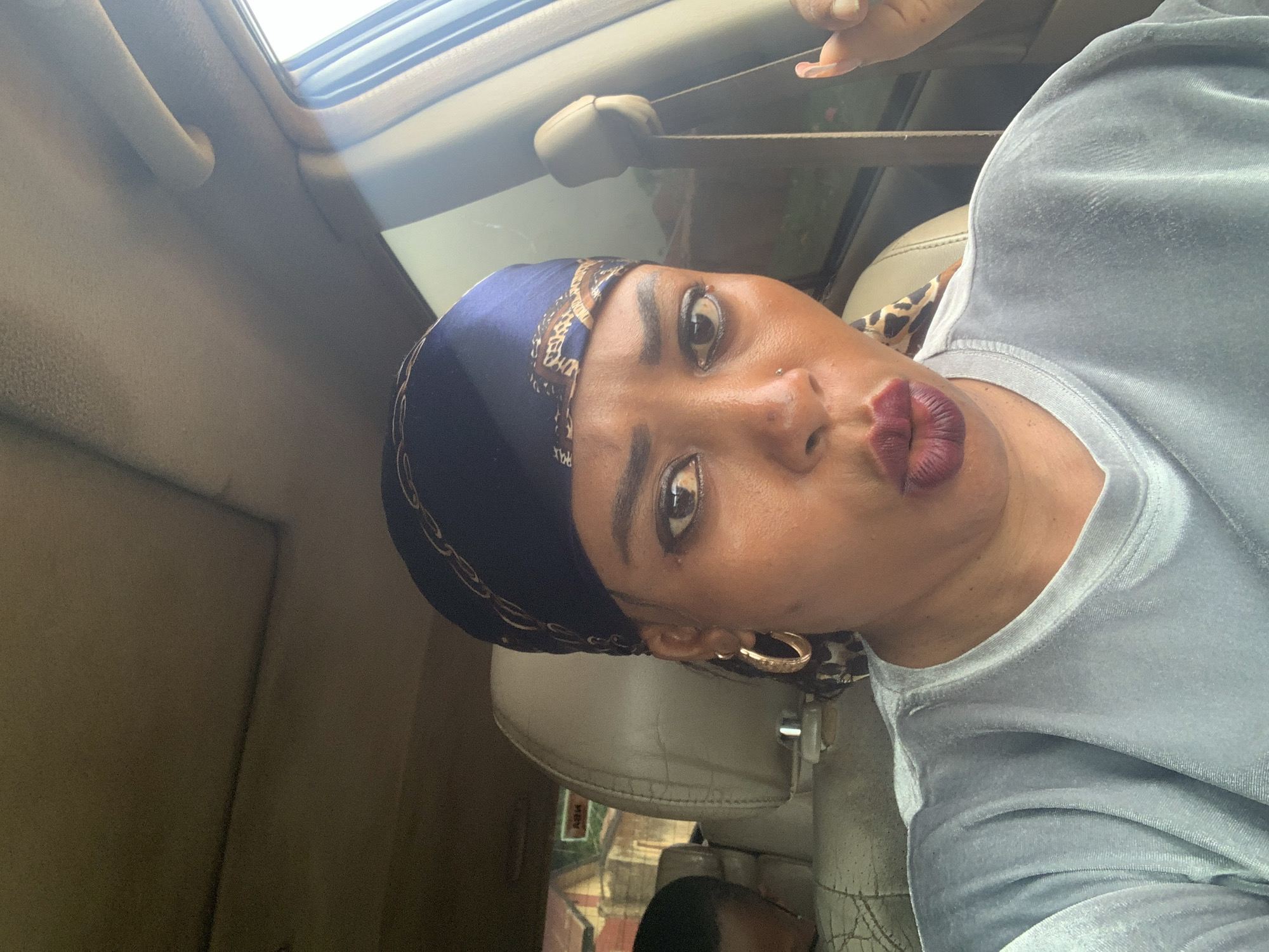 A woman is taking a selfie inside a car in Sabon Tasha, Nigeria. She is wearing a blue headscarf with a gold pattern, large gold hoop earrings, and a light gray velvet shirt. Her lips are pursed in a kiss. The interior of the car has beige-colored upholstery and a sunroof.
