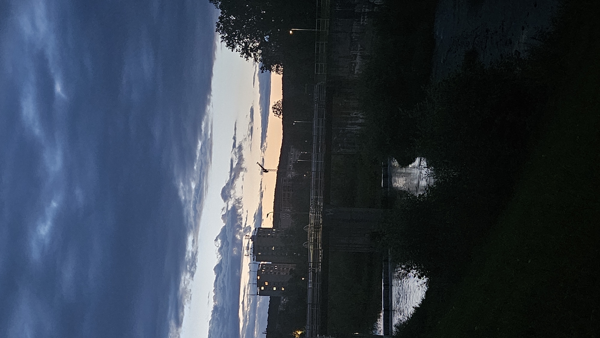 The image captures a dusk scene in Prešov, Slovakia, likely near the Torysa River. A concrete bridge spans across the water, with visible structural supports. The river flows beneath, with some rippling water suggesting a gentle current or a small weir. On the far bank, dense vegetation and trees create a dark silhouette. In the background, the city skyline is visible, featuring two prominent apartment buildings with illuminated windows, indicating evening activity. A construction crane stands taller than the buildings, silhouetted against the twilight sky. The sky itself is dramatic, dominated by heavy, dark clouds with lighter streaks of color near the horizon, hinting at a sunset or the approach of night. Streetlights are present on the bridge, casting faint glows. The overall atmosphere is serene yet slightly ominous due to the dark clouds.