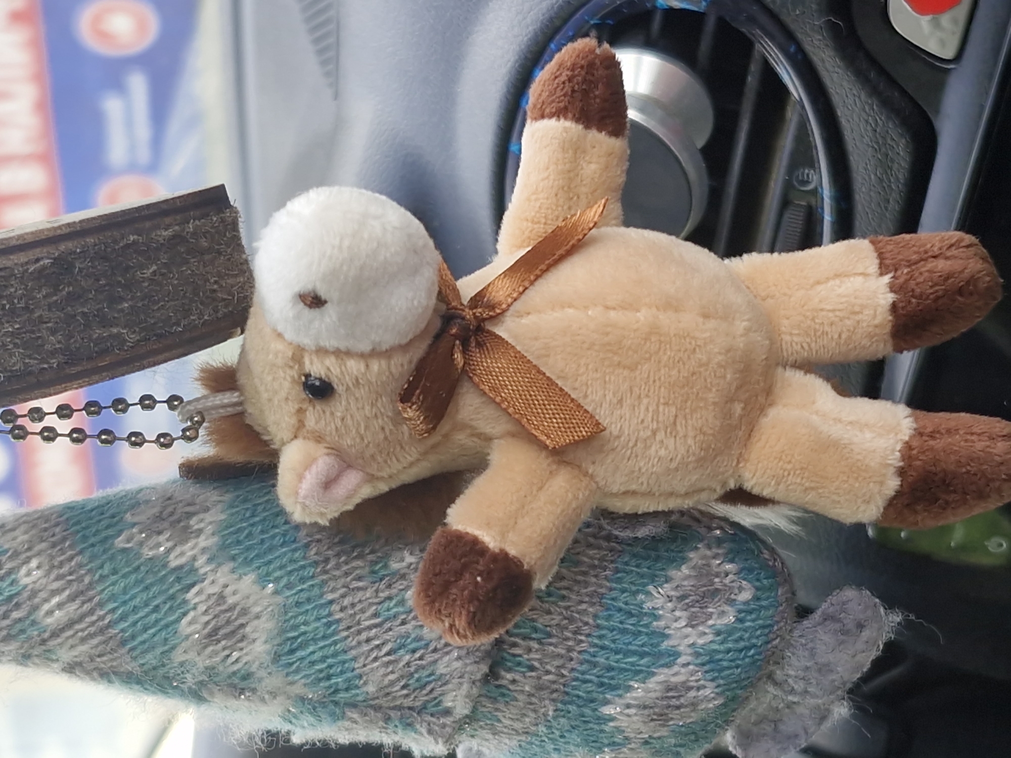 A stuffed toy animal, resembling a horse, rests on a fuzzy, patterned steering wheel cover inside of a vehicle. The toy is light brown with darker brown patches on its hooves and the tip of its tail, and it has a white patch on its head. A brown ribbon is tied around its middle. A small wooden object hangs from the toy with a beaded chain. Part of the dashboard and air vents are visible behind the toy. The location of Chaikovsky, Russia is not evident from this image.