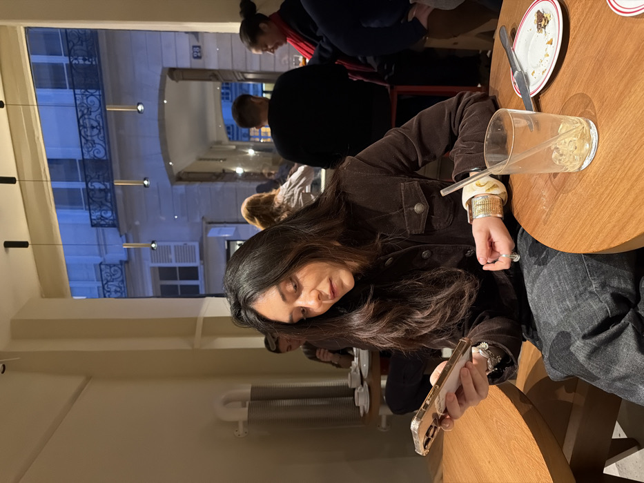 The image portrays an indoor scene, likely a cafe or restaurant in Paris, featuring a woman seated at a wooden table with an urban street visible through a large archway in the background. The main subject, a woman with long dark hair, wears a dark brown jacket and dark jeans. She is engrossed in her smartphone, held in both hands, which has a light-colored, possibly clear, case. Her left wrist displays multiple bracelets, including a wide, light-patterned one and several thinner metallic bands, and she wears rings on her fingers.
On the round wooden table before her, an empty or nearly empty glass with a straw and a small plate with cake remnants and a fork are present. Several other blurred individuals are visible in the background, seated at other tables, including one person wearing a red scarf.
Beyond the archway, typical Parisian architecture with wrought-iron balconies and stone buildings is discernable. A faint number "84" is visible on a building facade. The dark exterior suggests the time of day is evening or night, illuminated by artificial streetlights and interior pendant lights. A white radiator is also visible on an indoor wall. The overall impression is one of relaxed contemplation, with the woman focused on her device in a tranquil urban setting.