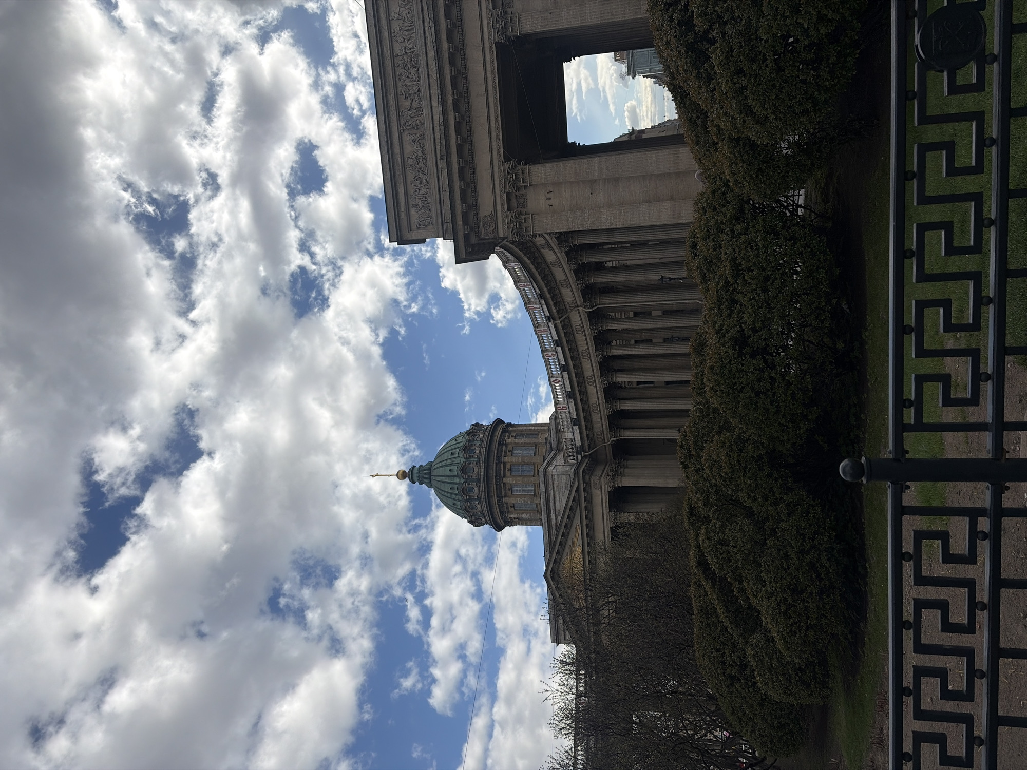 The image is an upward-angled, outdoor shot featuring the Kazan Cathedral in Saint Petersburg, Russia, framed against a sky with a mix of blue patches and puffy white clouds. The Cathedral's dome, topped with a gold spire, is visible. Part of the building's colonnade is visible as well. A portion of an ornate black metal fence with a geometric pattern is present in the right foreground, partially obscuring the view of greenery behind it. The perspective suggests the photo was taken from ground level.