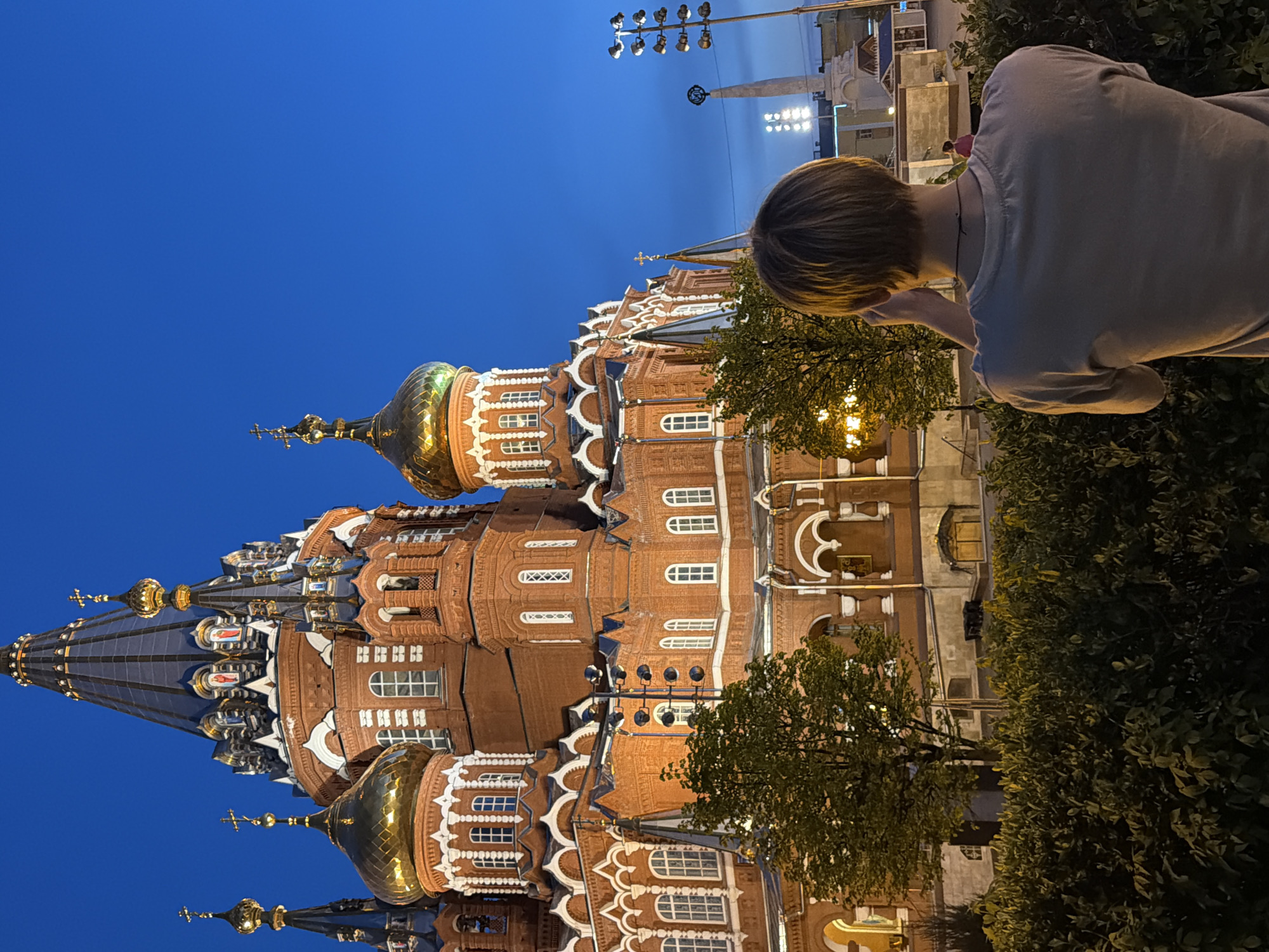 A low-angle shot depicts the Cathedral of Archangel Michael in Izhevsk, Russia, captured at dusk. The cathedral, constructed from red brick, showcases intricate architectural details, including arched windows, white decorative trim, and multiple golden and blue domes topped with crosses. The sky above is a deep blue, typical of twilight. A person with short blond hair, facing the cathedral, is positioned in the right side of the frame, possibly taking a photograph. The person wears a gray t-shirt. Bushes and trees are in the foreground. Street lights are in the background. The cathedral is illuminated, highlighting its facade and the golden domes.