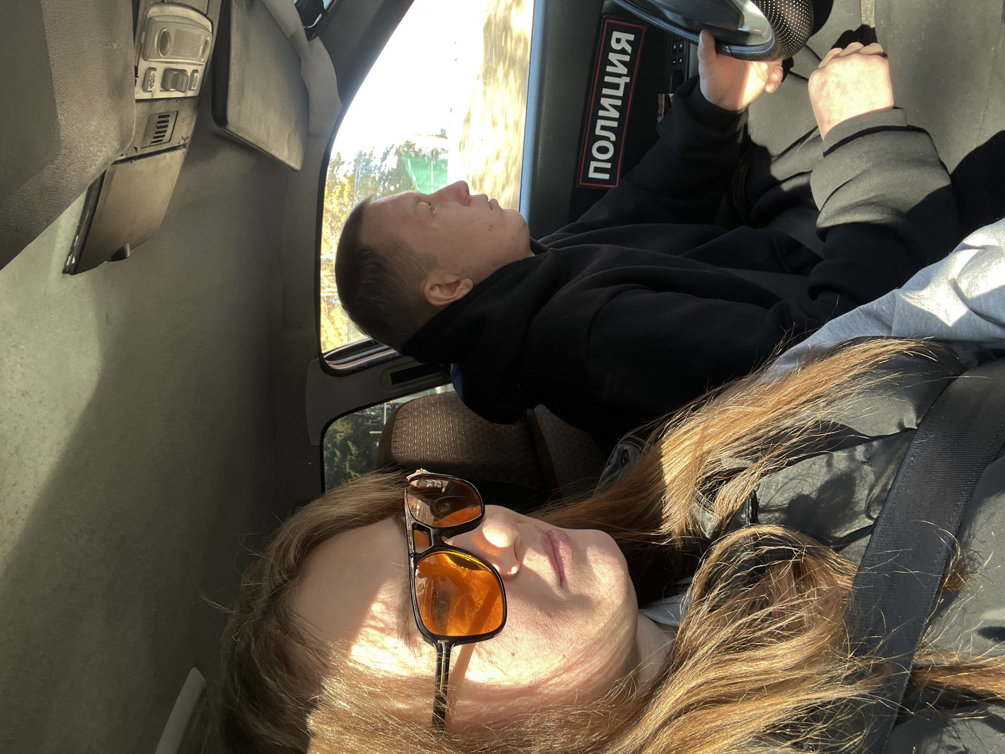 The image depicts the interior of a vehicle, presumed to be in Chaikovsky, Russia, during daylight hours with strong sunlight. In the foreground on the left, a female passenger with long, reddish-brown hair wears dark-framed sunglasses with orange-tinted lenses, a dark jacket, and a visible black seatbelt across her chest. Her face is illuminated by sunlight, and she gazes forward and slightly to her right. To her right, a male driver is visible in profile, wearing a black hooded sweatshirt. His hands are on the steering wheel, which has a dotted cover. A rectangular sign or patch displaying the Cyrillic text "ПОЛИЦИЯ" (POLICE) in white letters on a red and black background is affixed to the vehicle's interior near the steering wheel, likely on the dashboard or door panel. Through the vehicle's windows, green foliage and a bright, clear sky are visible. Overhead, the vehicle's grey headliner, a sun visor, and an overhead light fixture are discernible.