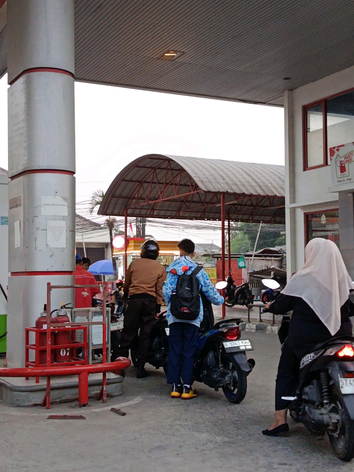 At a gas station in Bekasi, Indonesia, two individuals stand on motorcycles, one of whom is being attended to by a gas station attendant. The attendant, wearing a brown jacket, appears to be interacting with the person on the motorcycle. Another individual, a young person wearing a blue and white patterned shirt and carrying a backpack, stands behind the first motorcycle. A third person, wearing a white hijab and black clothing, is seated on a motorcycle to the right, facing away from the camera. In the background, a red fire extinguisher is visible on a metal rack. The gas station features a covered canopy with a corrugated metal roof. The scene depicts a typical transaction at a fuel station.
