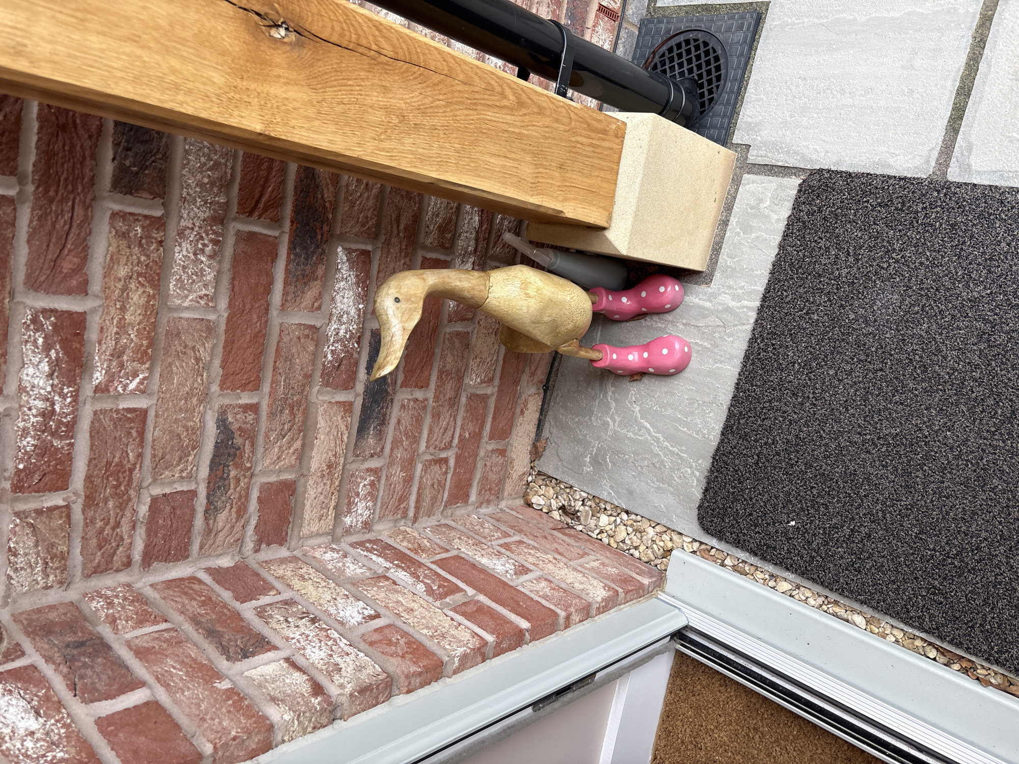 The scene shows a wooden duck decoration positioned on a brick ledge adjacent to a doorway in Longden, United Kingdom. The duck, carved from light-colored wood, is positioned so its head angles down. Two pink painted feet with white polka dots extend horizontally from the duck's body. Above the duck is a wooden beam with a dark-colored pipe attached. The ledge is constructed of red and white bricks. To the right of the ledge, there are gray paving stones, a dark gray mat, and a section of the door frame. Small stones are visible between the brick ledge and paving stones.