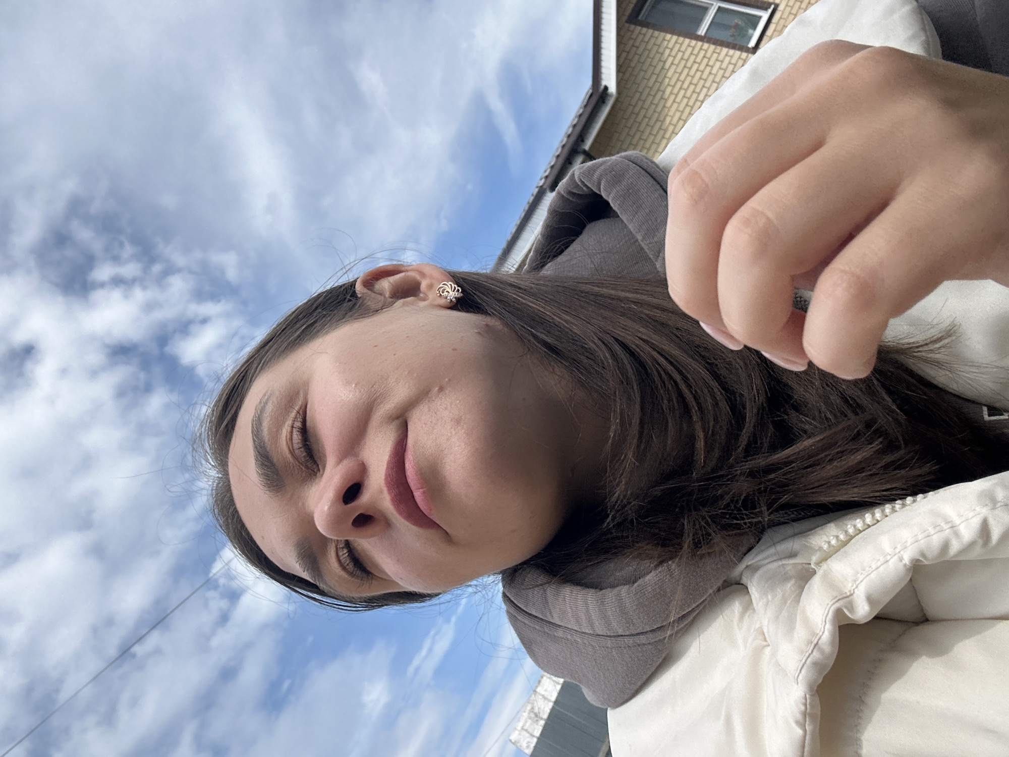 The image shows a low-angle, close-up shot of a young woman outdoors. She is wearing a light-colored puffer jacket over a gray hooded sweatshirt. Her eyes are closed, and she is looking upwards, with her head tilted back slightly. Her right hand is raised near her face, with her fingers gently touching her hair. She is wearing a small, intricate earring in her visible right ear.
The background consists of a bright blue sky with scattered white clouds. A portion of a brick building with a brown roof and a window is visible on the right side of the frame. A thin wire is also visible in the sky.
The setting appears to be residential, possibly a suburban area. While the exact location within Chaikovsky, Russia, is not discernible from the image alone, the visual cues (architecture, sky, general environment) are consistent with a Russian location. The lighting suggests it is daytime, likely under overcast or partly cloudy conditions. There are no other people visible in the frame. The overall mood is one of peaceful contemplation or enjoyment of the outdoor environment.