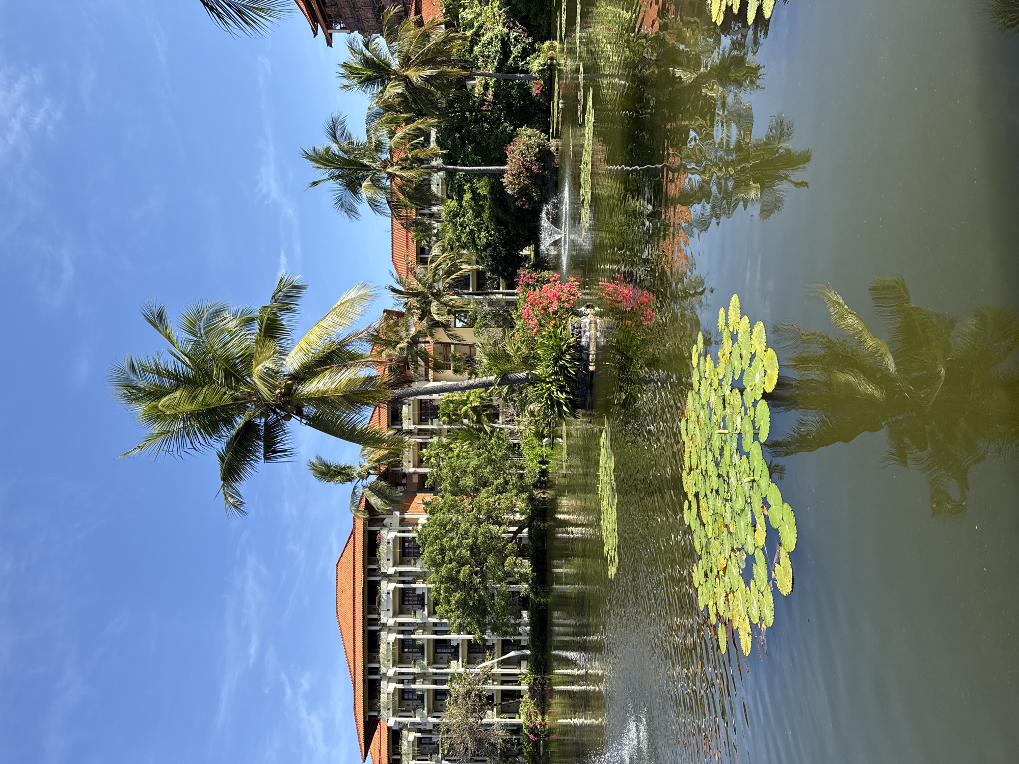 The image depicts a serene landscape in Kuta Selatan, Indonesia. A multi-story building with a red-tiled roof stands amidst lush greenery. Palm trees dominate the scene, with one prominent tree in the foreground. The building and trees are reflected in the calm water of a pond or lake, which also features lily pads. A small fountain is visible in the water, adding a touch of dynamism. The sky is clear blue, suggesting a bright, sunny day.