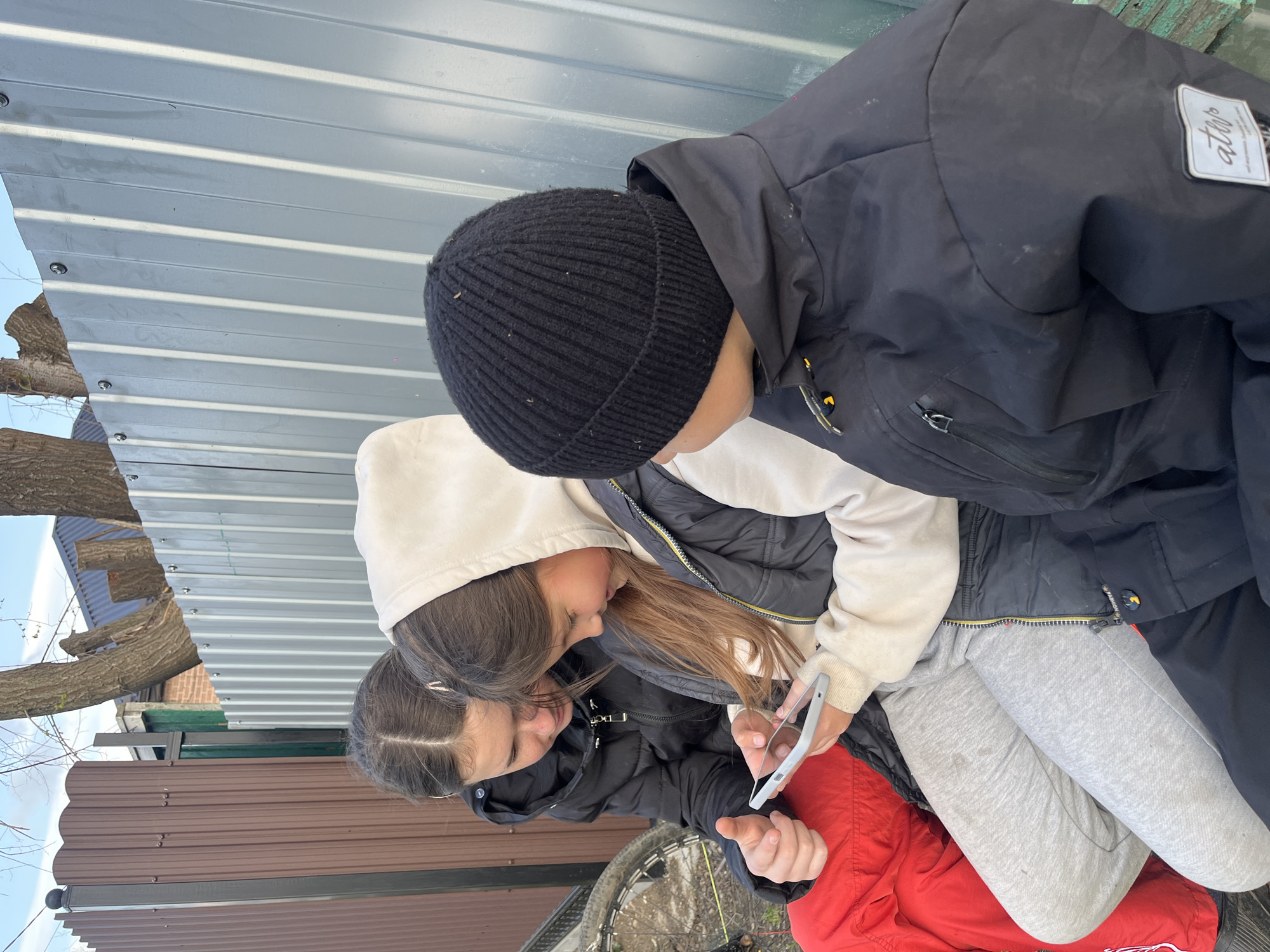 Two children are looking at a phone. One child wears a black beanie and a black jacket. The other wears a white hoodie and a black jacket. The child with the white hoodie sits on the lap of someone wearing gray sweatpants, who is sitting on someone wearing a red jacket. A corrugated metal fence and a brown metal fence are visible in the background. A tree branch is visible in the upper left. The location is implied to be in Novopokrovskaya, Russia.