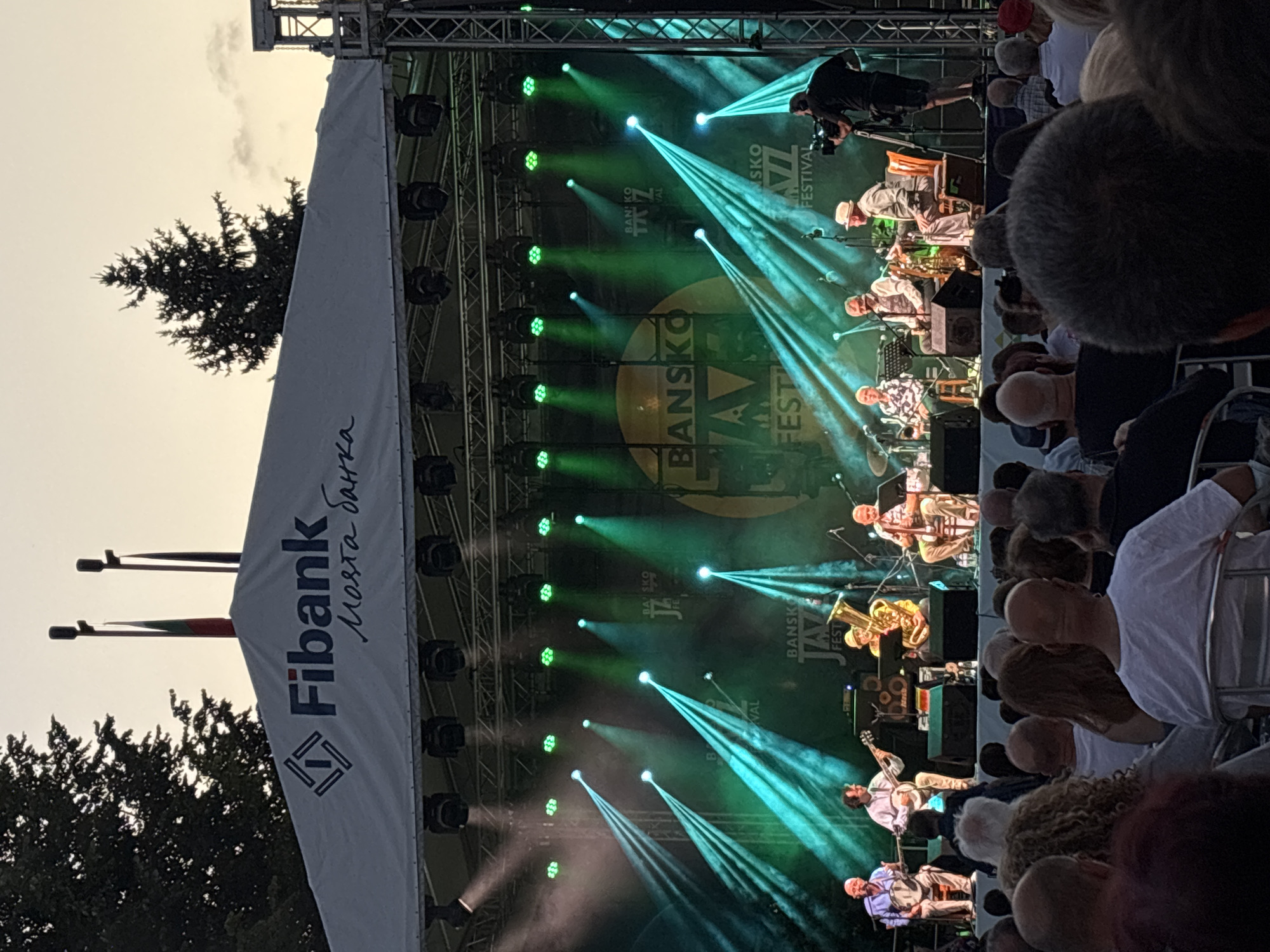 The image shows an outdoor jazz festival in Bansko, Bulgaria, possibly during a summer evening. A band of five musicians is performing on a stage under a large white canopy. The canopy has the "Fibank" logo and Bulgarian text on it. The stage is illuminated by numerous green spotlights creating beams of light that cut through the smoky air. The band members are seated and playing various instruments, including what appears to be a banjo, a guitar, a tuba, and a clarinet. The audience is seated in chairs in front of the stage, with their backs to the camera, suggesting a wide shot of the event. Some members of the audience are looking towards the stage, while others are engaged in conversation. A cameraman is filming the performance from the right side of the stage. The background shows trees and a twilight sky. The overall atmosphere seems relaxed and festive.