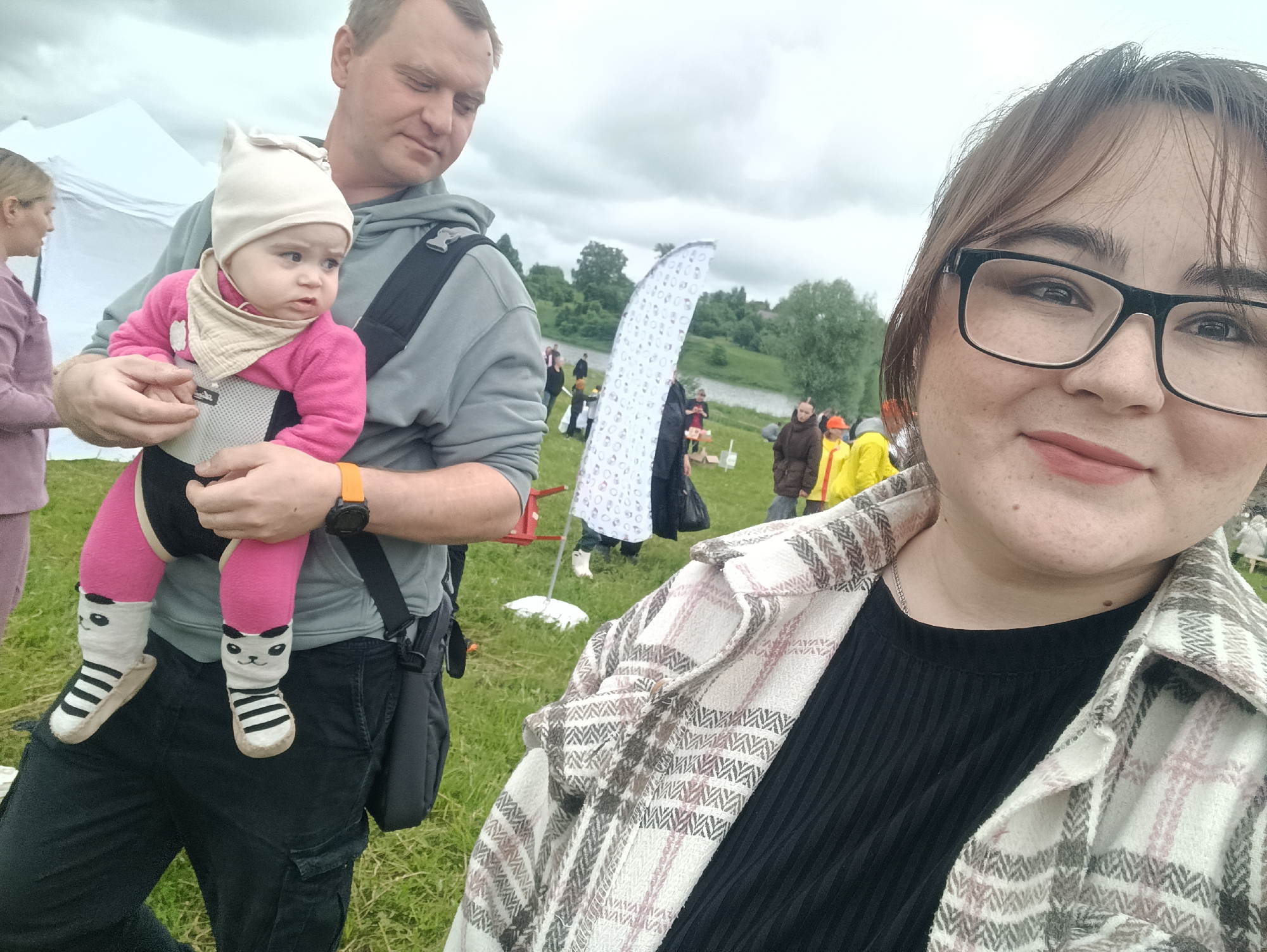 This outdoor photograph depicts a group of people at an event on an overcast day in Unknown, Unknown. In the immediate foreground, a woman with brown hair and black-framed glasses is taking a selfie, smiling slightly towards the camera. She wears a plaid jacket featuring white, grey, and light pink patterns over a black ribbed shirt.
To her left, in the mid-ground, a man with short brown hair holds a baby in a front-facing carrier. He wears a grey hoodie, black pants, and an orange watch on his left wrist. The baby, facing forward, wears a light beige pointed hat, a beige bandana bib, a bright pink long-sleeved outfit, and white socks with black stripes and cartoon panda faces on the feet. The baby's gaze is directed slightly to its right. Partially visible behind the man is another woman wearing a purple garment.
The background reveals a grassy field sloping down towards a body of water, likely a river or lake, bordered by trees. Several individuals are dispersed across the field; some are indistinct, while others are seen wearing items such as yellow raincoats or dark jackets. A large, curved, white structure with a repeating pattern stands upright in the mid-background. A white tent is visible on the far left. The sky is overcast with a uniform grey cloud cover.