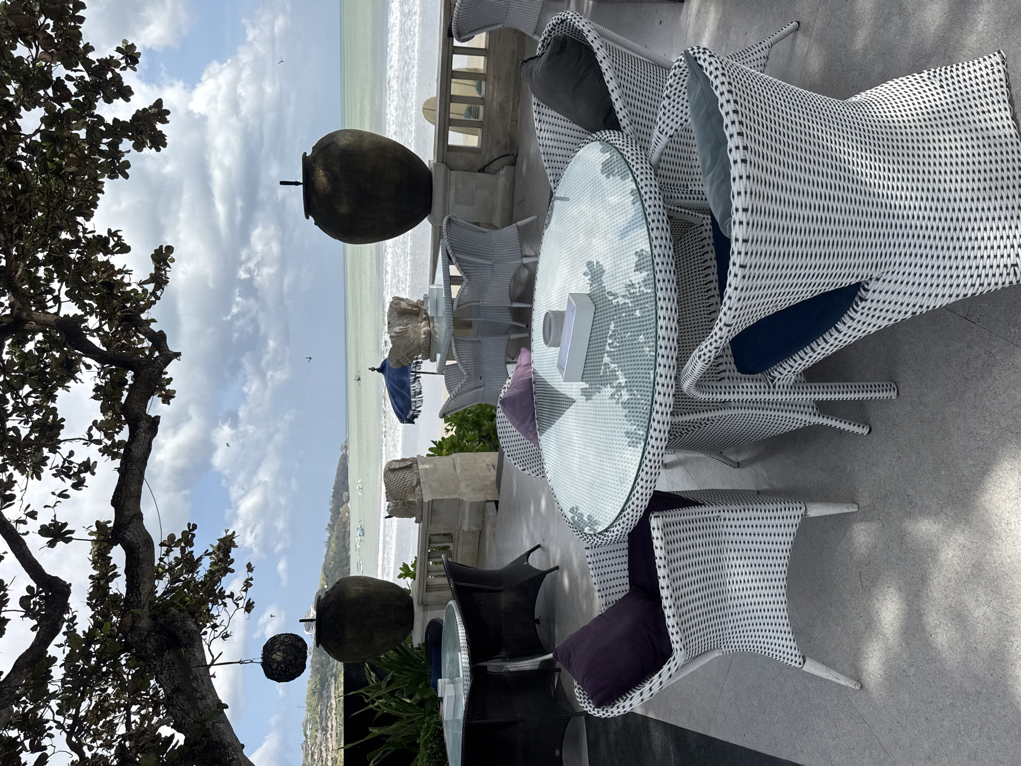 A patio dining area overlooks a beach in Kuta Selatan, Indonesia. The foreground features a round glass-top table with several white wicker chairs surrounding it. One chair has a dark grey cushion and a dark purple pillow. Another chair has a dark blue cushion and a dark grey pillow. A white rectangular box rests on the table. In the background, a larger rectangular dark wicker table is set with chairs. Beyond the patio, a railing separates it from the beach. The sea is visible with gentle waves breaking on the shore. Several boats are anchored in the water. The sky is partly cloudy. Large decorative vases are placed on the patio, and a tree with dark green leaves overhangs the scene.