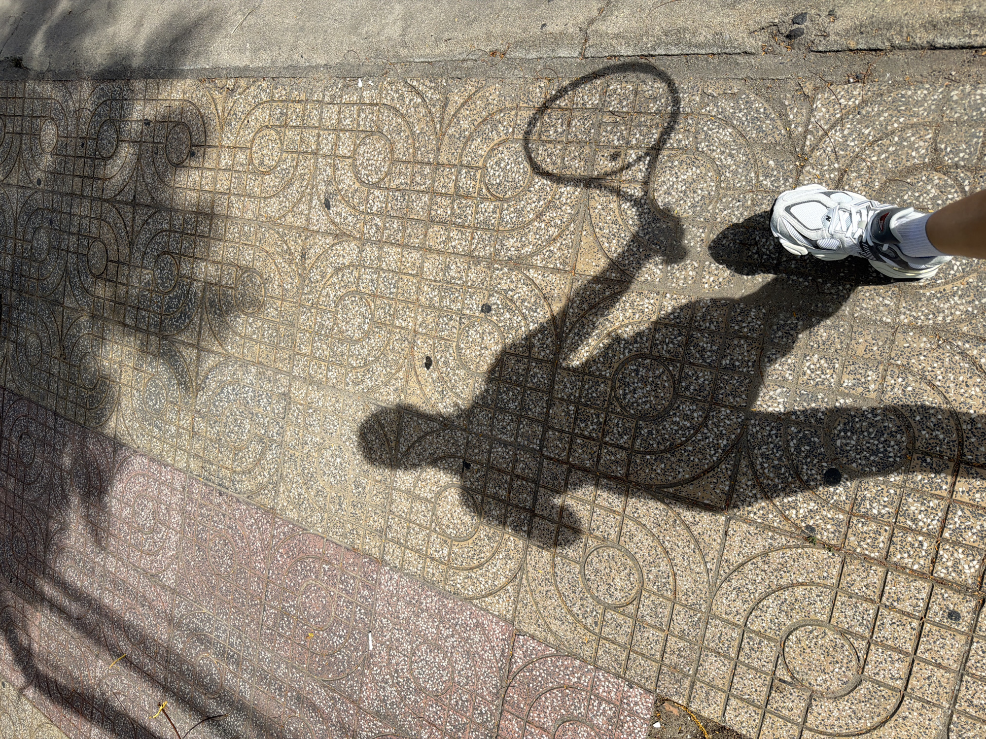 A person's shadow is cast on a tiled sidewalk in Bac Nha Trang Ward, Vietnam. The shadow depicts the person holding a tennis racket and wearing athletic shoes. The sidewalk is made of square tiles with circular patterns. Sunlight casts tree shadows on the pavement. The scene suggests an outdoor activity in a urban setting.