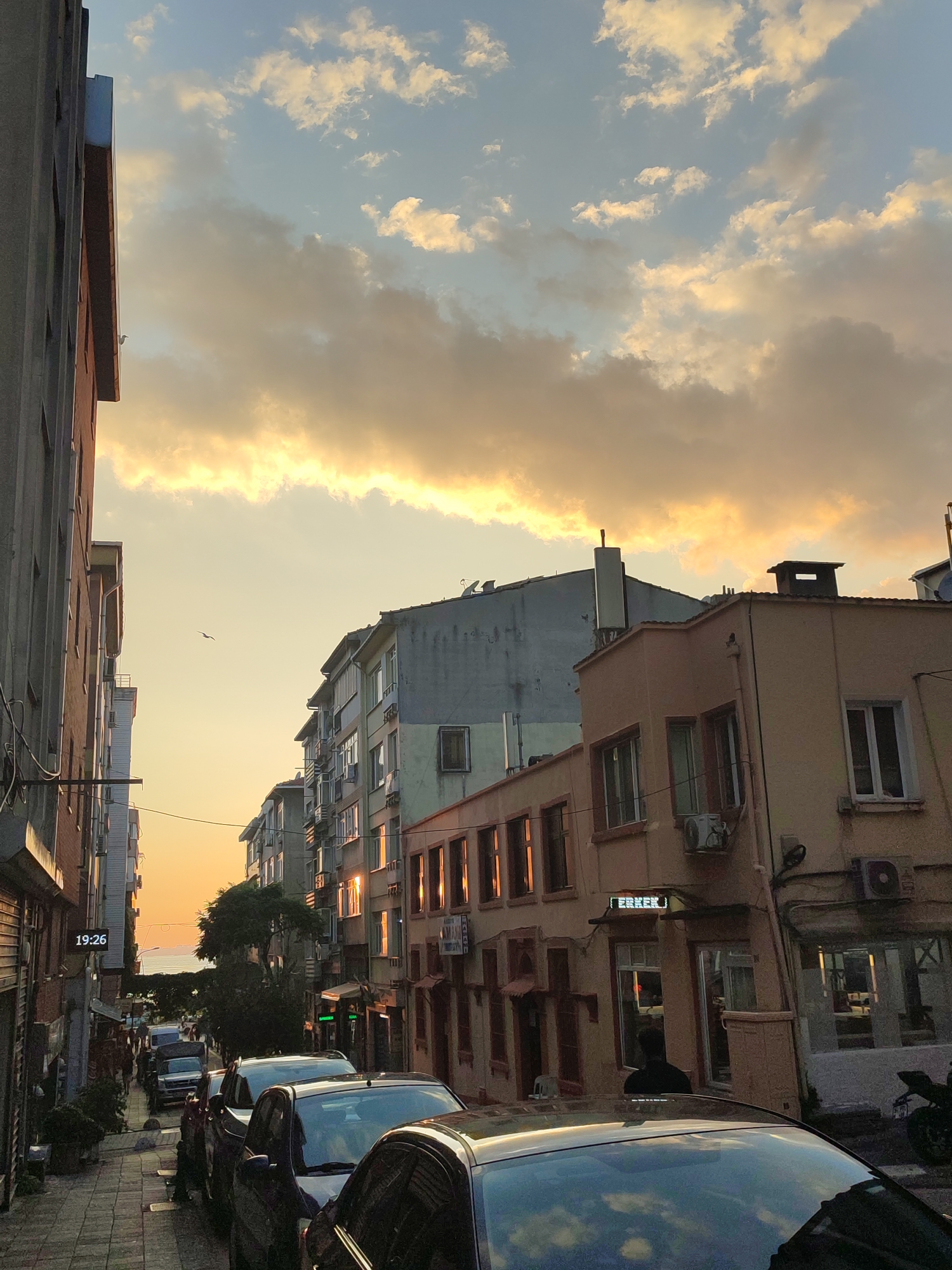 The image depicts a narrow street in Kadıköy, Turkey, bathed in the warm glow of sunset. Buildings line both sides of the street, their facades reflecting the orange and yellow hues of the sky. Several parked cars are visible, occupying much of the roadway. The sky is filled with dramatic clouds, tinged with the colors of sunset. A single bird can be seen in flight against the luminous sky. The overall impression is one of a quiet urban evening.