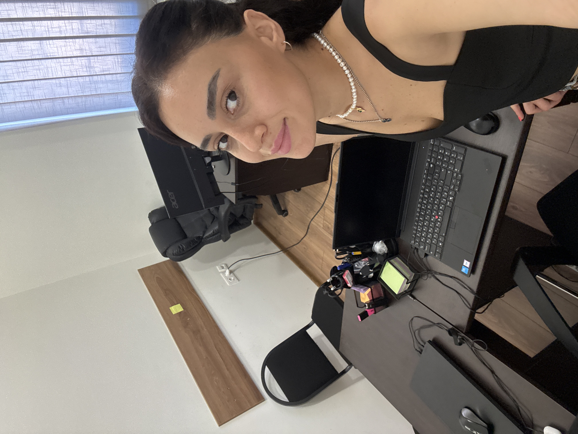A woman with dark hair, wearing a black top and a pearl necklace, smiles at the camera. She is in an office setting, with a dark wood desk in the foreground. On the desk, there is a laptop, a mouse, a black laptop stand, and office supplies in a black organizer. Behind her, a black office chair and a computer monitor are visible. The walls are white, and there is a wooden plank mounted on the wall. A window with vertical blinds is in the background. The image is taken from a slightly elevated angle.