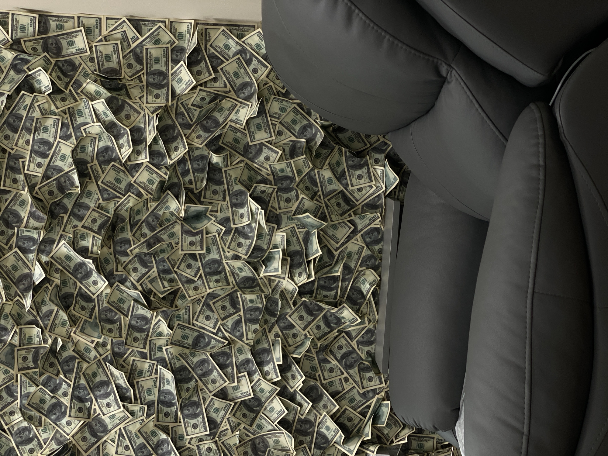 The image displays a floor covered in numerous U.S. one hundred dollar bills, juxtaposed against the corner of a dark grey, leather-like sofa. The dollar bills are scattered randomly, creating a thick layer on the surface. The sofa occupies the right side of the frame, its sleek material contrasting with the disarray of money on the floor. The location is not deducible from the visual information.
