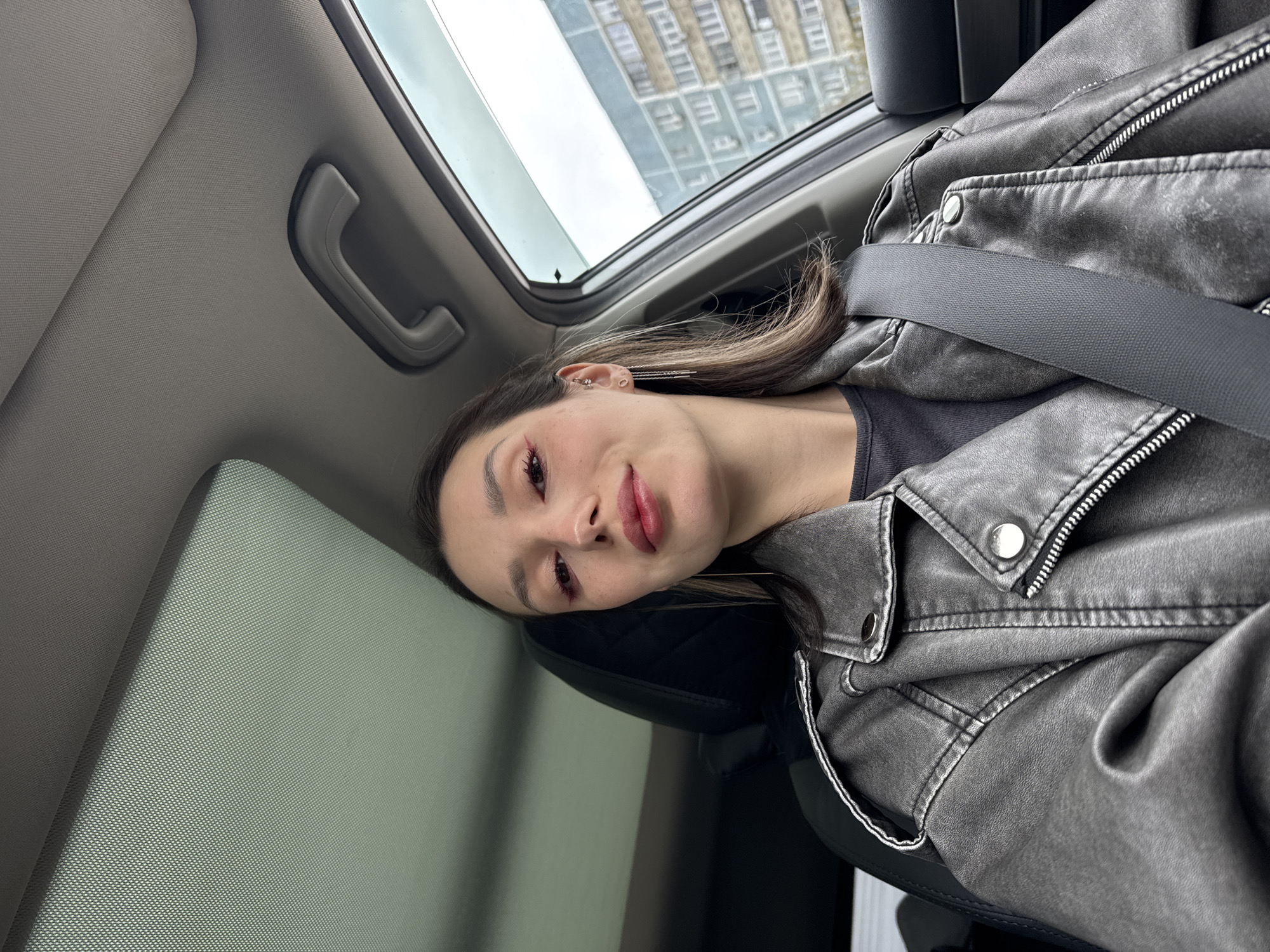 A woman is seated in a car, wearing a gray leather jacket and a seatbelt. The car has a sunroof. Outside the sunroof, a tall building is visible. The location appears to be an urban environment.