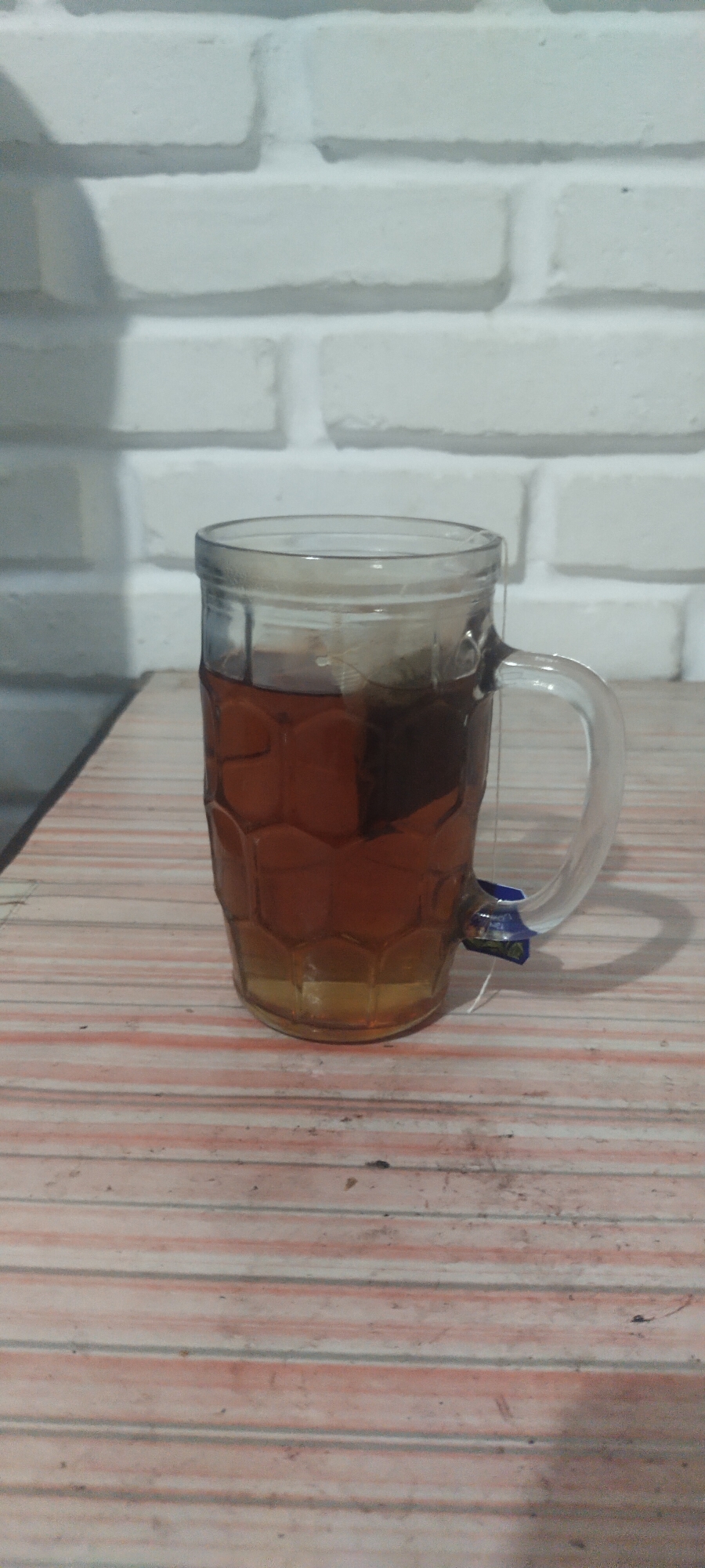 A clear glass mug filled with amber-colored tea, with a tea bag submerged within, sits on a wooden table. The table has a striped pattern of white, pink, and faint gray lines. Behind the mug, a white brick wall serves as the background. The scene appears to be indoors. There are no people present in the image. The overall impression is a simple, still-life composition. The location is specified as Pasirhuni, Indonesia, however, there are no visual cues in the image to support this specific location.