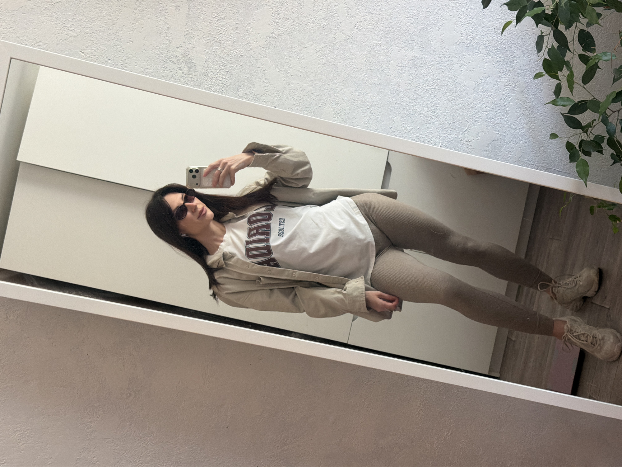 The image is a mirror selfie of a woman in Perm, Russia. She is wearing sunglasses, a white t-shirt with a maroon graphic, a beige button-down shirt, beige leggings, and beige sneakers. The mirror is rectangular with a white frame. The background includes a white wall, a white closet, a potted plant, and a hardwood floor. The woman is holding a smartphone. The location is likely an apartment or house.