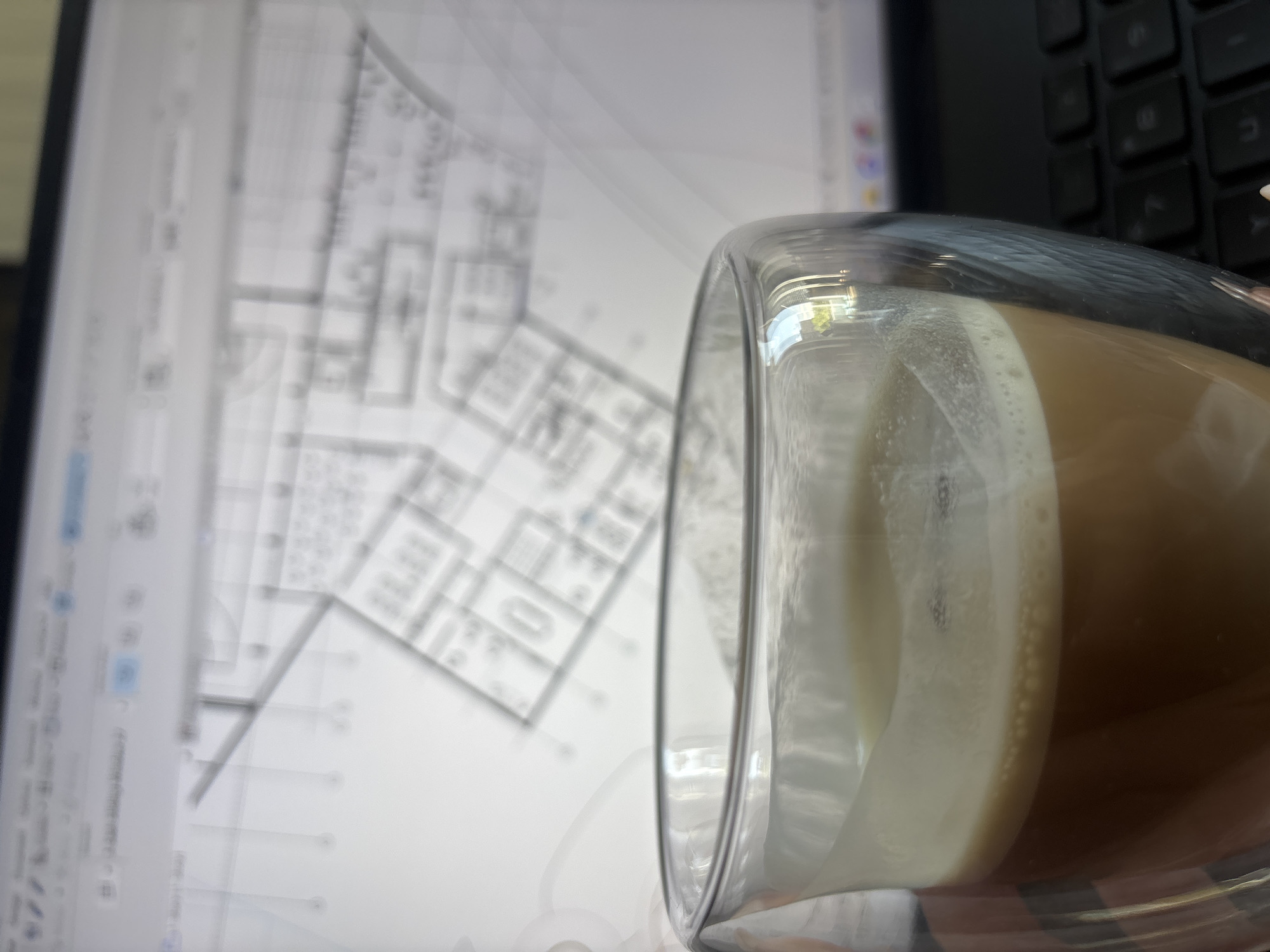 The image shows a close-up of a glass of coffee with milk, with an architectural blueprint displayed on a computer screen in the background. The computer is next to a black keyboard, partially visible. The focus is on the glass and its contents, with the blueprint and keyboard blurred. There is no information to determine the location to be Yerevan, Armenia.