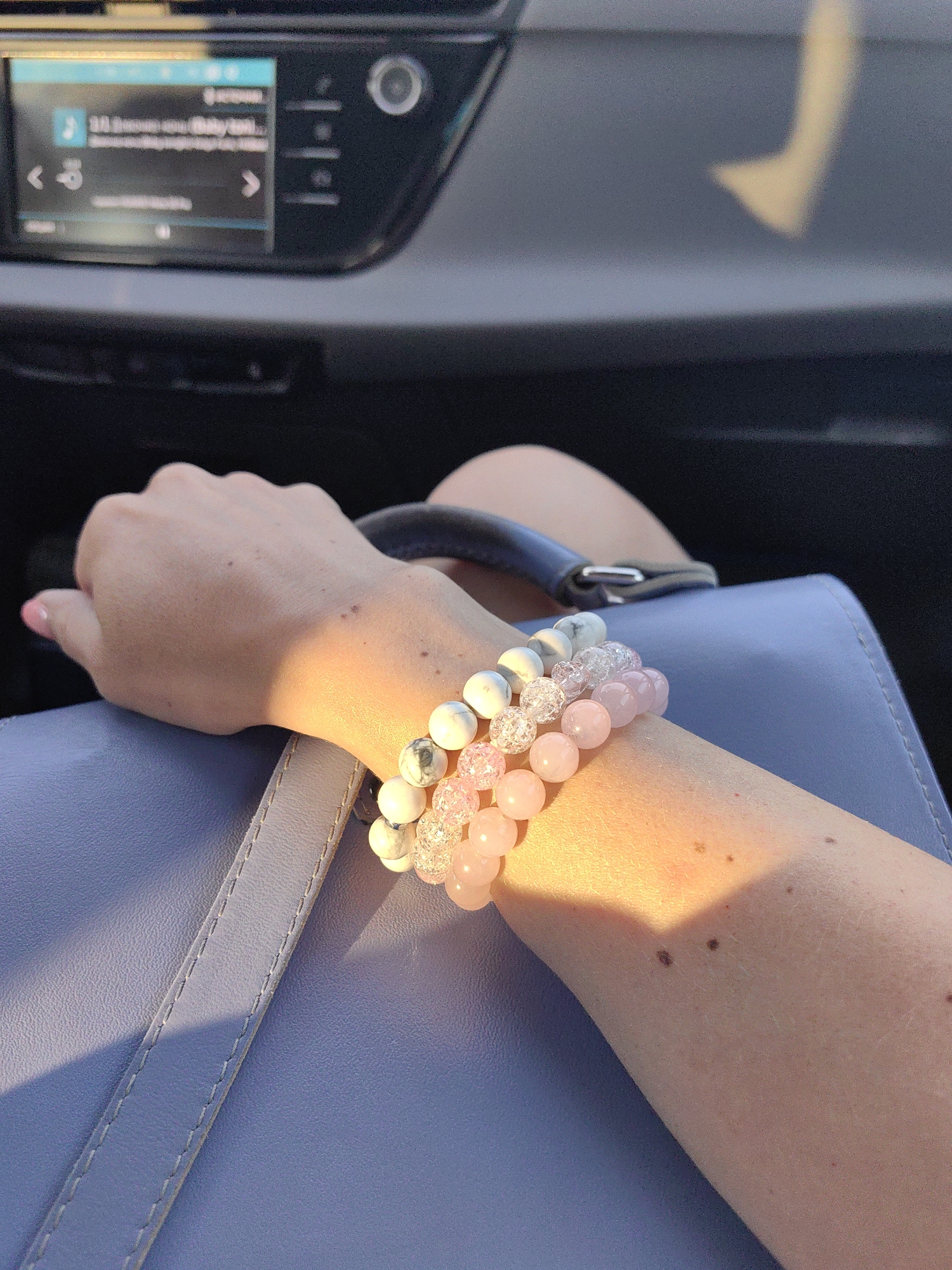The image shows a close-up of a person's arm wearing three beaded bracelets. The bracelets consist of white and grey marbled beads, clear textured beads, and light pink rose quartz beads. The arm is resting on a light blue textured surface, possibly a bag or purse, and a dark gray steering wheel is partially visible in the background. Sunlight is casting a warm glow on the arm and bracelets. The background includes a car's dashboard with a media display. The location is identified as Лугаваслабадскі сельскі Савет, Belarus.
