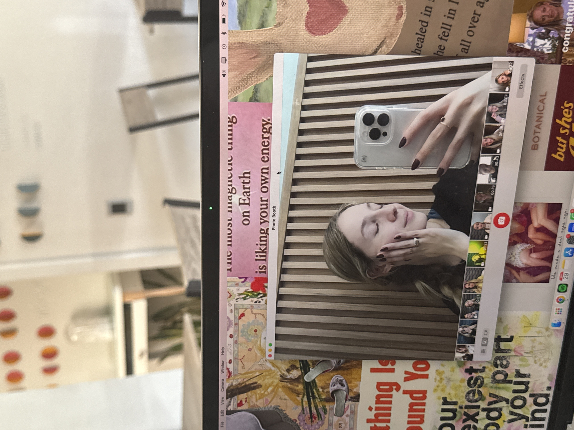 The image captures a laptop screen displaying a vibrant digital collage, rotated 90 degrees clockwise, against a softly blurred indoor background in Mexico City, Mexico.
On the laptop screen, a prominent Photo Booth application window shows a woman with light brown hair, dark fingernail polish, and a ring on her finger. She is captured with her eyes closed, touching her cheek, while holding a modern smartphone (an iPhone with three visible cameras) which reflects her face. To the right of this main image, several smaller video thumbnails of the same woman are arranged vertically, with timestamps such as "00:02", "03:07", and "00:43", and an "Effects" button is visible. A green light, indicating active camera use, is visible near the top left of the screen.
The Photo Booth window is surrounded by a patchwork of text and graphical elements, forming a personal mood board or journal. Visible text includes empowering phrases like "the most magnetic thing on Earth is liking your own energy," and fragments such as "healed in," "BOTANICAL," "but she's," and "congratula." Another section reads "thing Is und Yo our exiest ody part your ind." A small red heart design is also part of the collage.
The macOS menu bar is visible along the top edge of the screen (appearing on the left due to the rotation), displaying "File Edit View Camera Window Help" and system icons for Wi-Fi, battery level, Bluetooth, and volume. The macOS dock is partially visible along the bottom edge (appearing on the right), showing icons for various applications and the date "29".
The background, out of focus, reveals a bright indoor setting with hints of modern furniture, possibly chairs or tables with dark frames, and decorative objects with circular orange patterns on a shelf to the left. The overall impression is one of personal reflection and digital creativity.