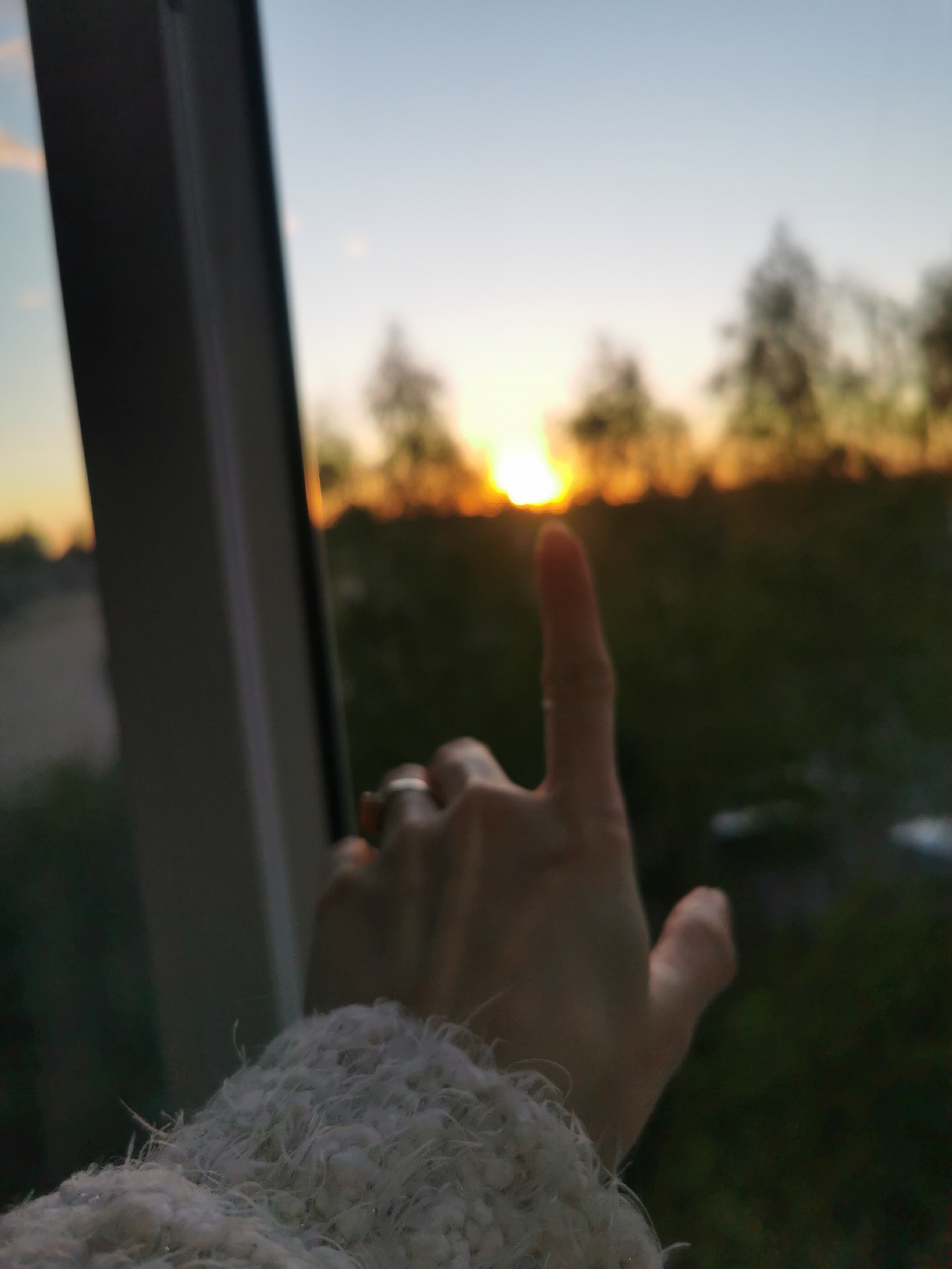 A blurry, soft-focus, medium shot depicts a hand pointing towards the sunset, seemingly taken from inside a building, possibly in Chaikovsky, Russia. A window frame is visible on the left, and the hand is positioned against a blurred background of trees and a field illuminated by the sunset. The person is wearing a white, textured sweater. A ring is visible on the ring finger of the hand. The location is inferred from the sunset scenery being common in Chaikovsky, Russia.