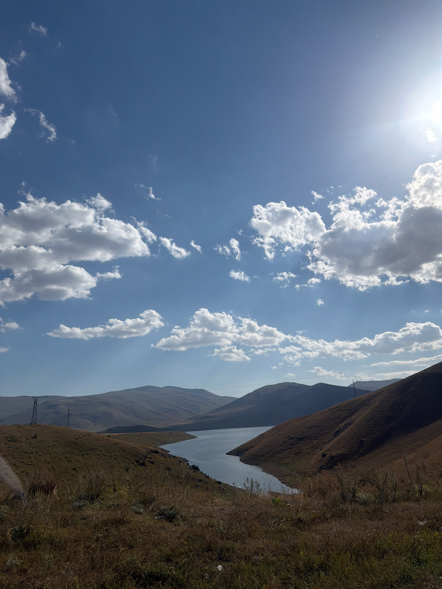 The image captures a panoramic view of a lake nestled within rolling, dry, grassy hills under a partly cloudy blue sky. The sun is visible in the upper right corner, casting a bright glare and some lens flare. The terrain is dominated by shades of brown and yellow from the dry vegetation, with some areas of darker green indicating sparse growth.
In the foreground, the dry, textured grass is the most prominent element, with some taller stalks and smaller, lighter-colored plants interspersed. The landscape recedes into the distance, showing the curved shoreline of the lake. The water itself appears calm and reflects the sky.
Further back, the hills rise and fall, their slopes etched with shadows that emphasize their contours. In the mid-ground and background, the outlines of more distant mountains are visible, appearing softer and more muted in color due to atmospheric haze.
Human-made structures are present but not central to the scene. On the left side, a tall, lattice-like transmission tower stands on a hill. Another similar tower is visible in the far distance, and power lines can be discerned strung between poles across the hills.
The overall impression is one of a natural, somewhat arid landscape, possibly in the late summer or early autumn, with signs of human infrastructure present. The location is identified as Tsghuk, Armenia. There are no people visible in the image.