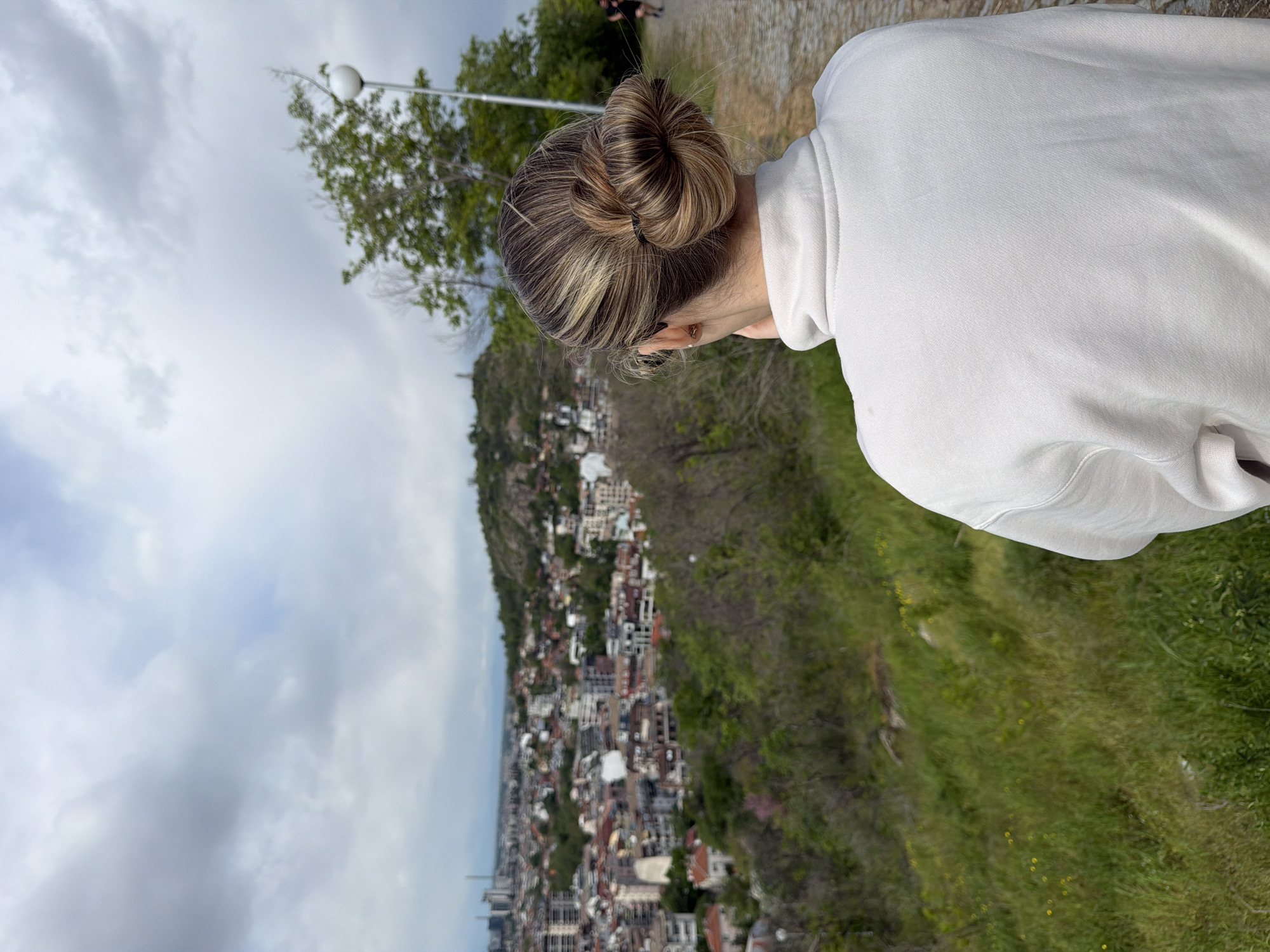 A woman wearing a white hooded sweatshirt stands overlooking the city of Plovdiv, Bulgaria. She has her back to the camera, revealing a hair bun. The city below is visible, with buildings and vegetation lining a hillside. A lamppost and trees are to the left of the frame. The sky is overcast.