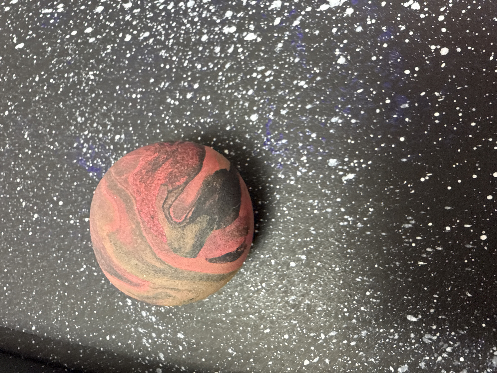 A close-up shows a speckled, dark gray surface with a mottled, egg-shaped object resting on it. The object features swirling patterns of red, tan, and black. The backdrop resembles a night sky with scattered white dots suggesting stars. The location, Chaikovsky, Russia, is not directly verifiable from the visual elements of the image alone.