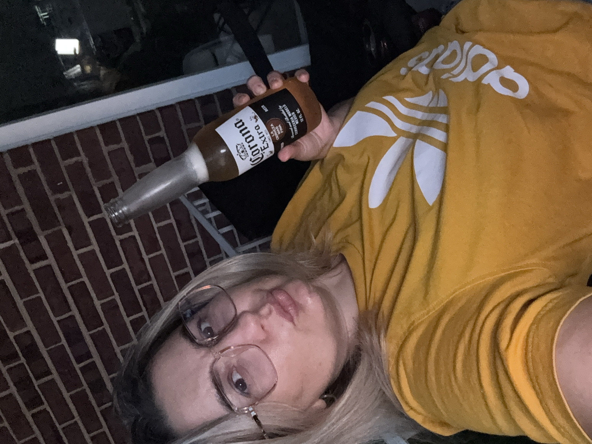 A woman with blonde hair and glasses is holding a bottle of Corona Extra beer. She is wearing a yellow t-shirt with an Adidas logo. The background appears to be outdoors, with a brick wall and dark shadows. The image is taken from a slightly elevated angle, focusing on the woman's face and the beer bottle. The lighting is dim, suggesting it may be evening or nighttime.