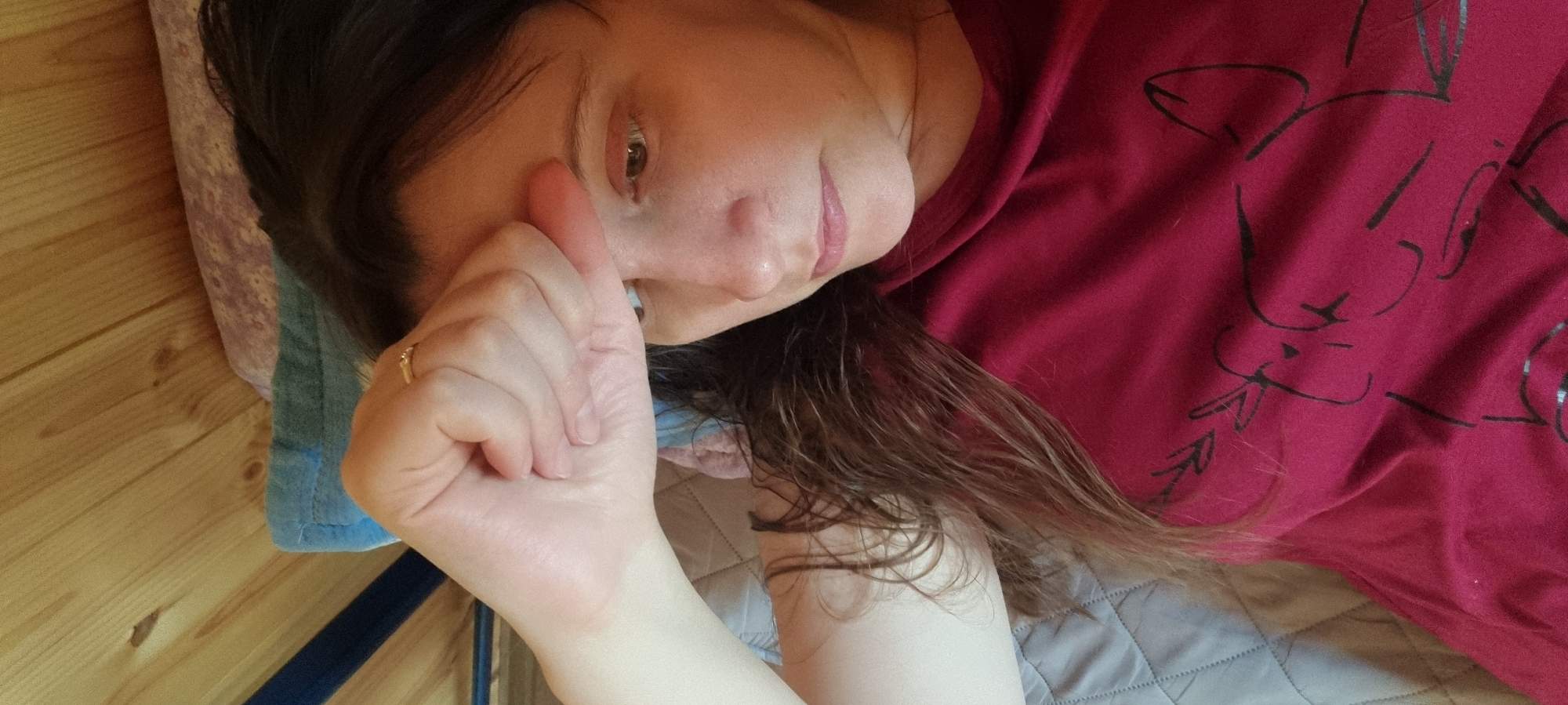 A woman is lying on her back, propped up on a pillow, in what appears to be an indoor setting. She is wearing a red t-shirt with a black design on it. Her left hand is raised towards her face, and she is wearing a ring on her left ring finger. Her hair is dark and slightly wet. The background includes a wooden floor and a grey quilted surface, presumably a bed or mattress. There is no explicit evidence to confirm this photo was taken in Dunnae-myeon, South Korea.