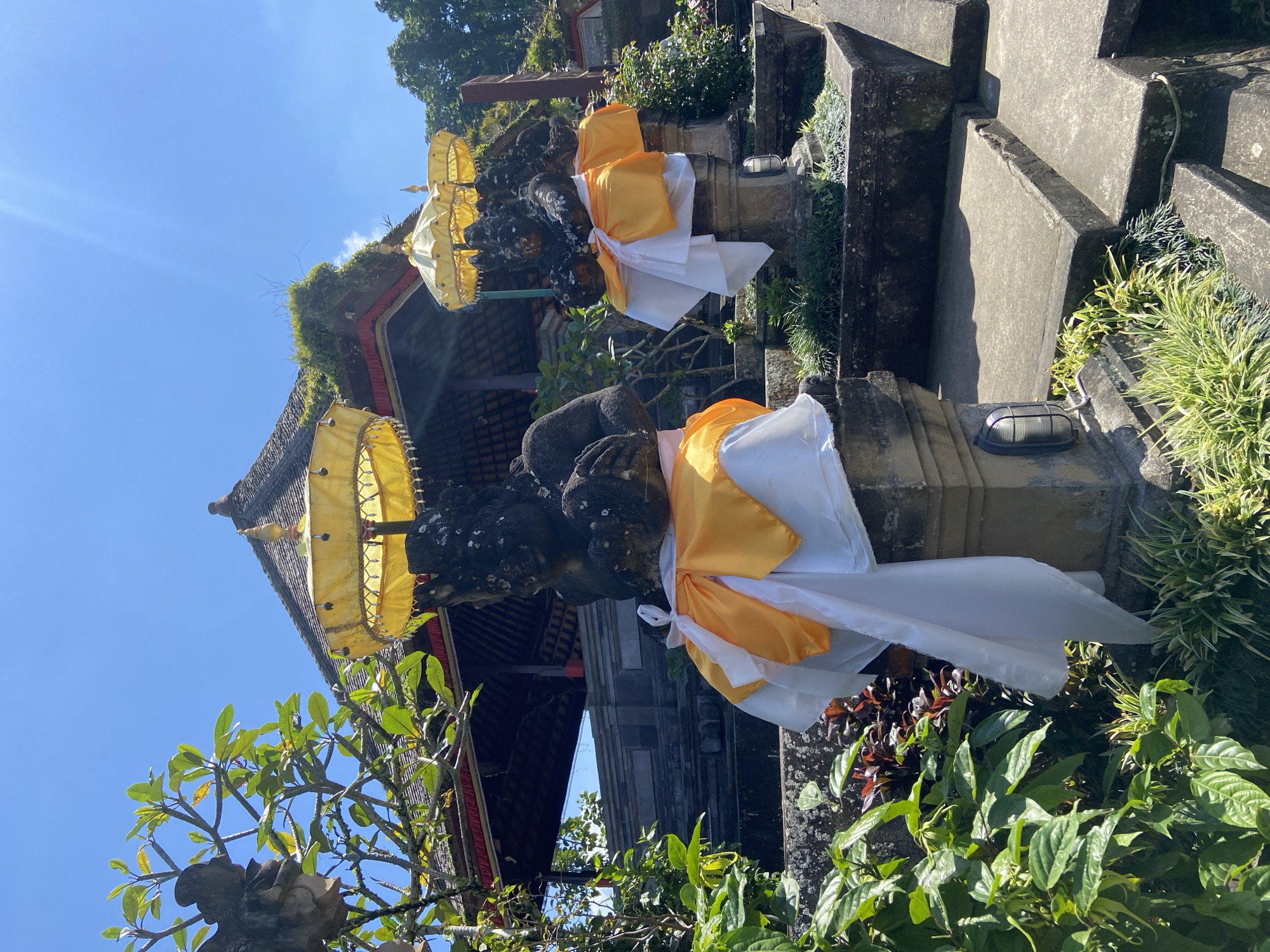 The image shows two stone statues, each adorned with yellow and white fabric, standing on pedestals in front of a temple structure. The statues appear to be guardian figures. The temple has a thatched roof and is surrounded by lush greenery. The sky is clear and blue, indicating a sunny day. The setting appears to be outdoors, likely in a temple complex. The location is the Pura Kehen temple in Bangli, Indonesia.