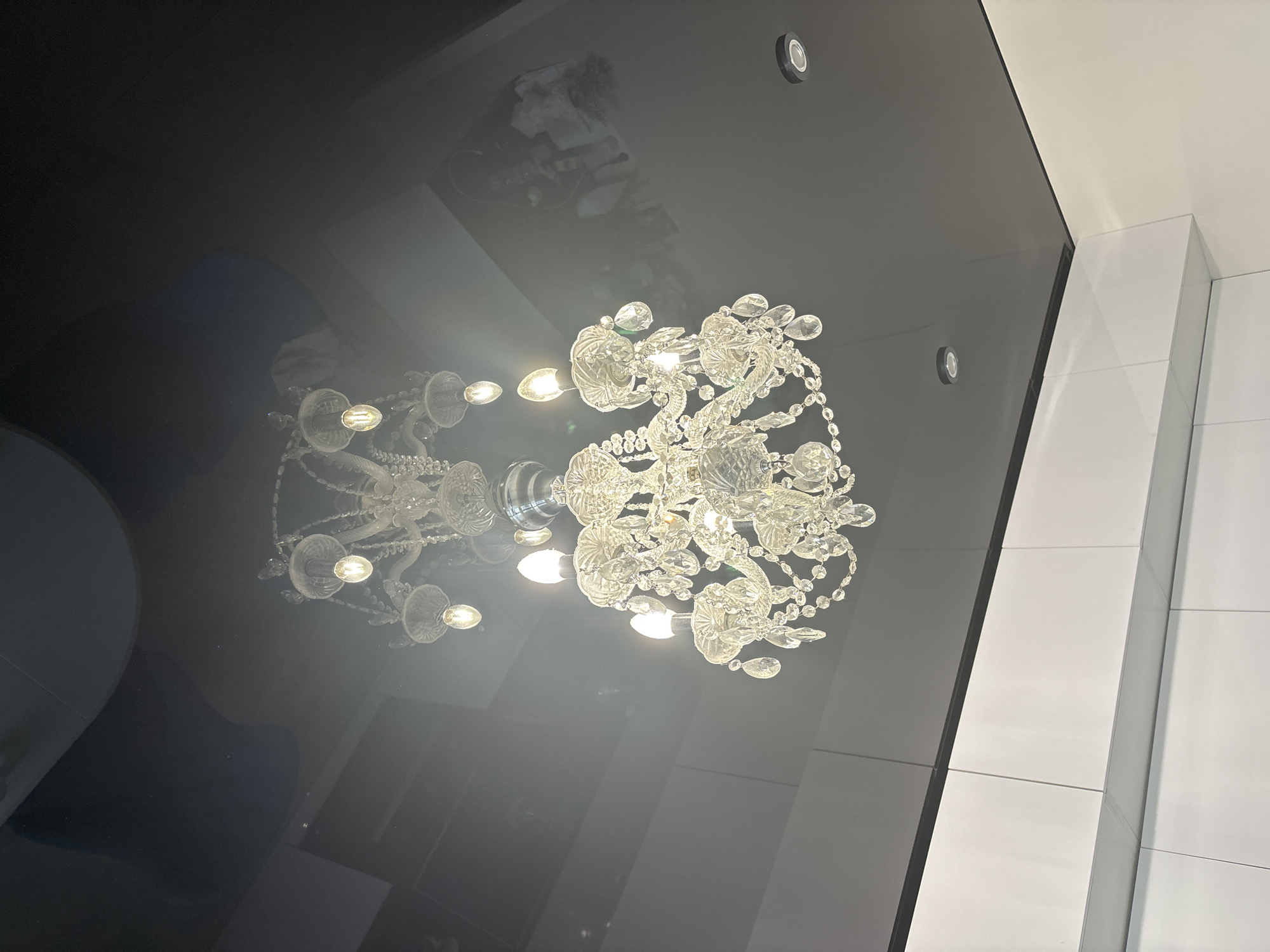 A low-angle shot depicts a crystal chandelier suspended from a glossy black ceiling, reflecting elements of the room. Adjacent to the ceiling is a white tiled wall. Recessed ceiling lights are visible. The location is Unknown, Unknown.