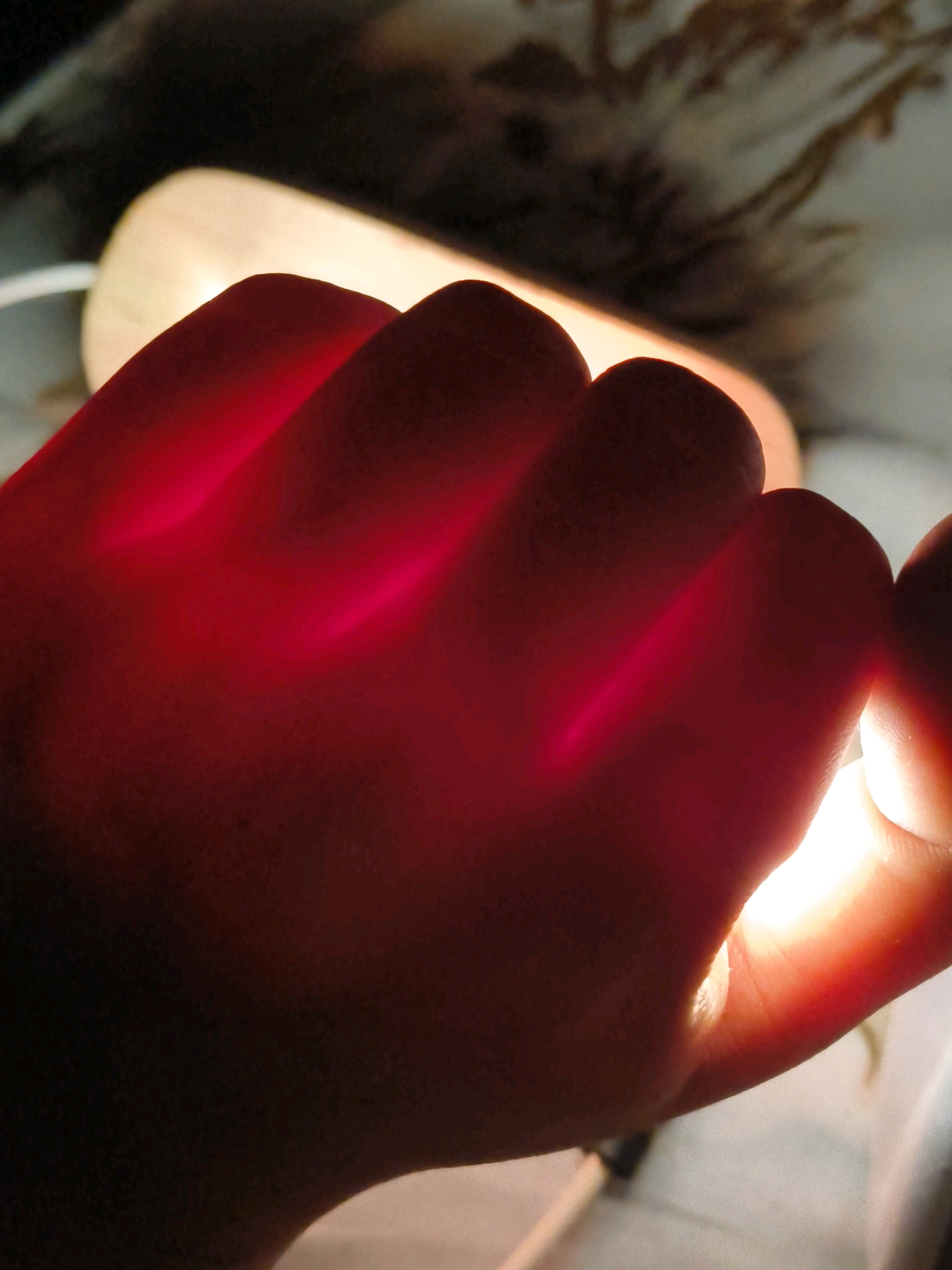 The image shows a close-up of a hand illuminated from behind by a red light source. The fingers are clenched into a fist, and the light shines through the skin, highlighting the contours of the knuckles and the spaces between the fingers. The background is blurred and appears to be a domestic environment with a light-colored surface and some indistinct objects, possibly textiles. There are no discernible elements that would allow for the identification of a specific location in Mohali, India, or any other geographical area. The scene is a static, focused shot on the hand and the play of light.