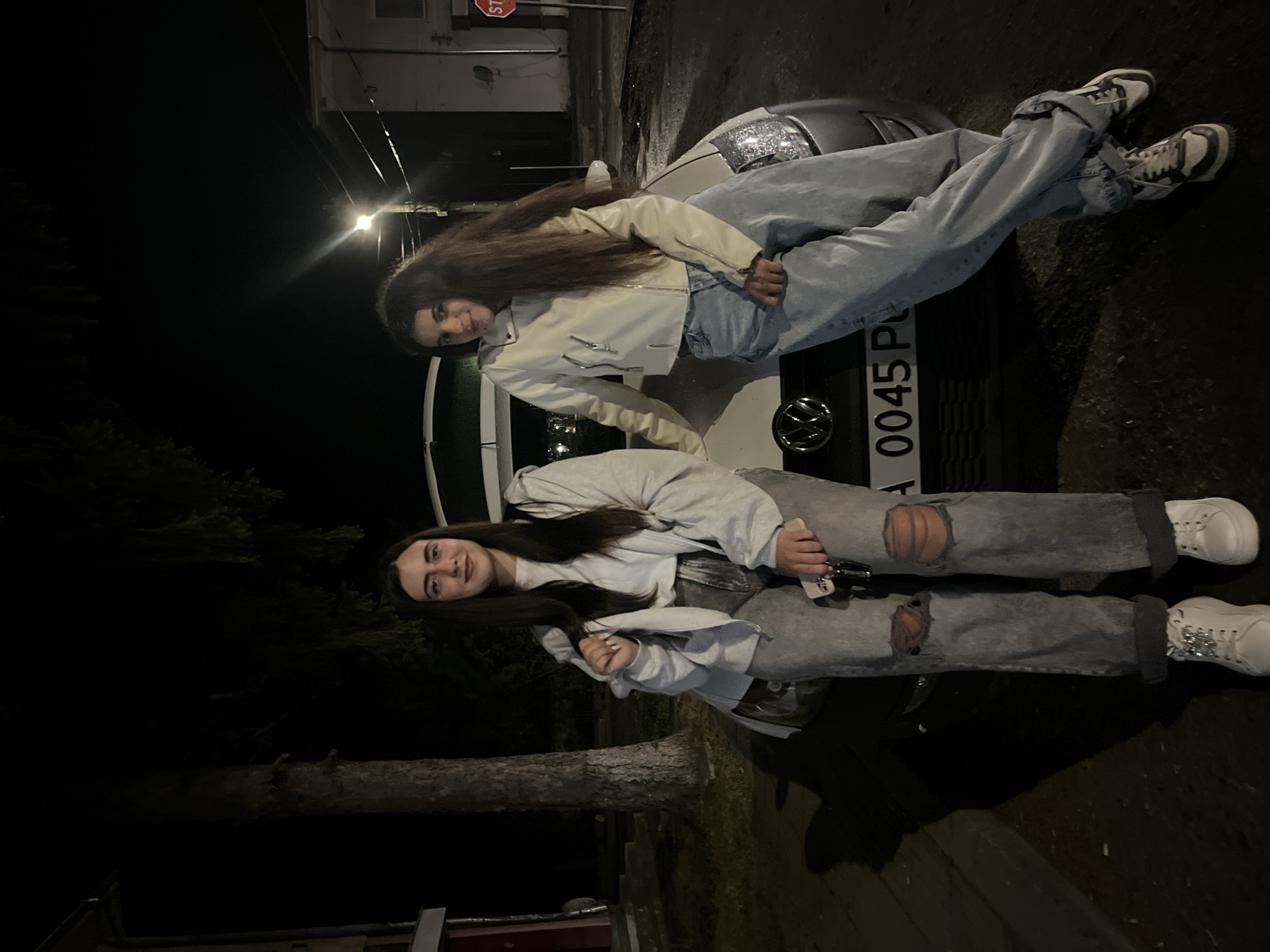 Two young women are posing on the hood of a parked silver Volkswagen car at night. The car has a license plate that reads "A 0045 P." The location appears to be Lozarevo, Bulgaria. One woman is wearing a white jacket and light blue jeans with dark shoes, she is sitting on the hood. The other woman is wearing a light grey hoodie and ripped grey jeans and white sneakers, she is in front of the car. A stop sign is visible. It is dark, illuminated by streetlights.