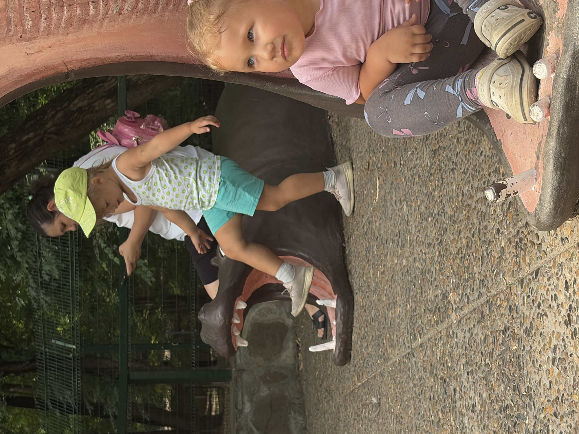 The image depicts an outdoor scene in Rostov-on-Don, Russia, likely in a public park or zoo. In the foreground, a young Caucasian girl with blonde hair tied in a ponytail sits on a large, stylized hippopotamus-shaped structure. She is wearing a pink t-shirt and grey leggings with a blue and pink bow pattern, and beige Croc-style shoes. Her gaze is directed upwards and to the right, suggesting she is watching something off-camera. Behind her, a second young Caucasian girl, also wearing a light-colored sleeveless top with green circles and turquoise shorts, is positioned on the hippopotamus sculpture, with one leg extended. She is wearing a light green cap and beige sneakers. A woman, partially visible behind the second girl, appears to be looking at her mobile phone. The hippopotamus sculpture is dark brown with a pink interior, resembling an open mouth with white teeth. The ground is a textured surface composed of small pebbles. In the background, there is a green metal fence, trees, and foliage, indicating a natural or semi-natural environment. The overall lighting suggests it is daytime, likely sunny.