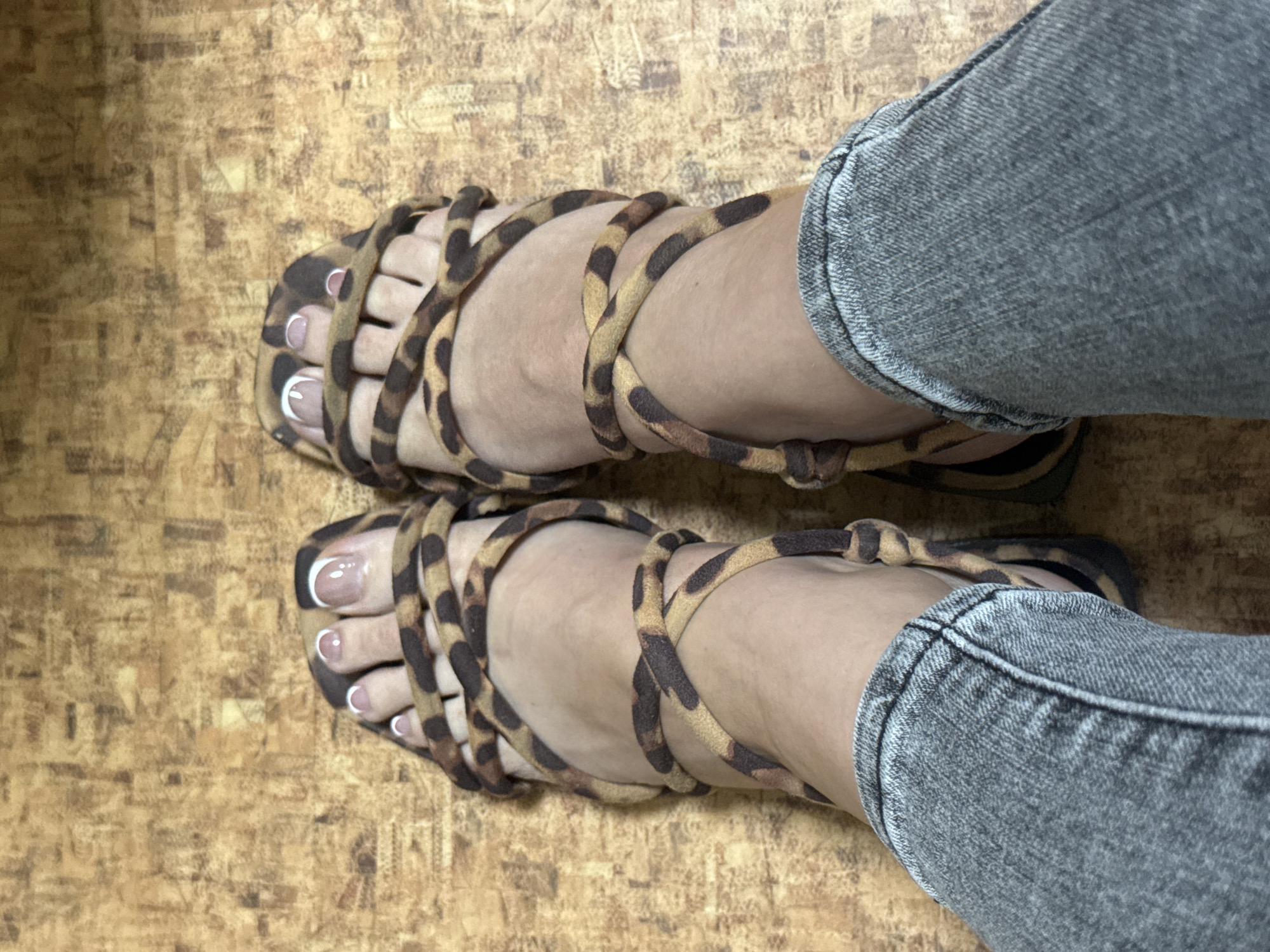 A pair of feet wearing leopard print lace-up sandals are shown from a high-angle perspective, standing on a wood-grain floor. The feet have French manicured toenails. The person is wearing grey denim jeans. The setting is indoors on a neutral floor.