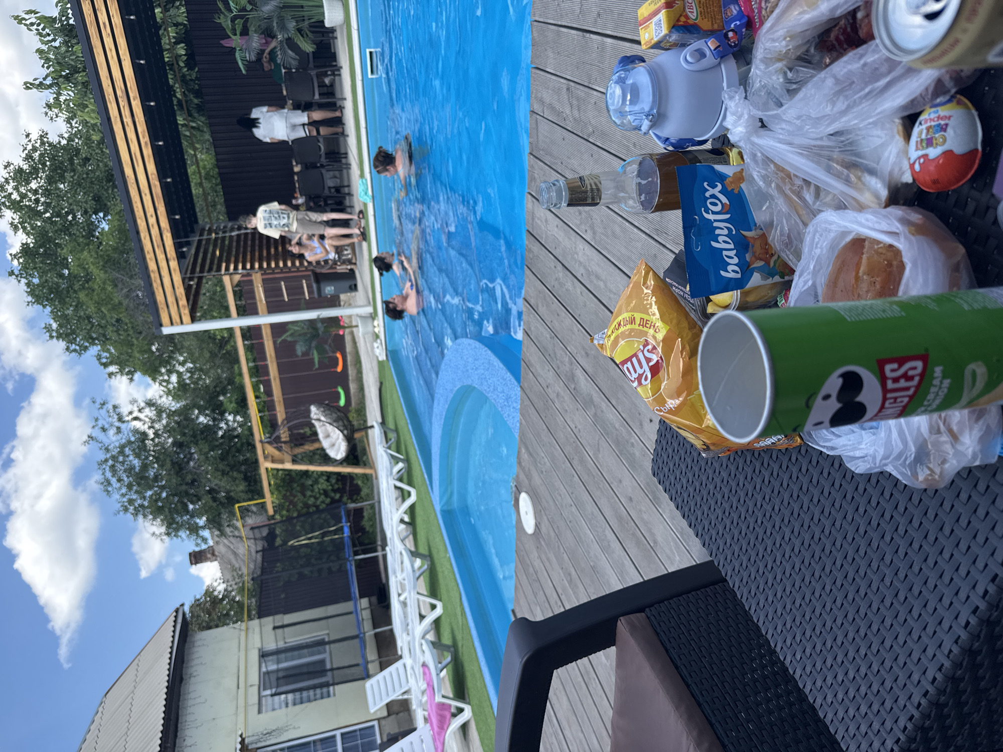 The image depicts an outdoor residential backyard scene with a swimming pool. In the foreground, a black wicker-style table holds various food items and drinks, including a can of Pringles, a bag of Lay's chips, a bottle of beer, a blue water bottle, a Kinder Surprise egg, and other snacks in plastic bags. Behind the table, a wooden deck leads to a vibrant blue swimming pool where several people are seen swimming and interacting. In the background, a house with a gray roof and white siding is visible, along with a trampoline and a swing set. Further back, a covered patio area with tables and chairs suggests a dining space. Lush green trees and a blue sky with white clouds complete the outdoor environment. The overall impression is a leisurely gathering by a pool on a sunny day.