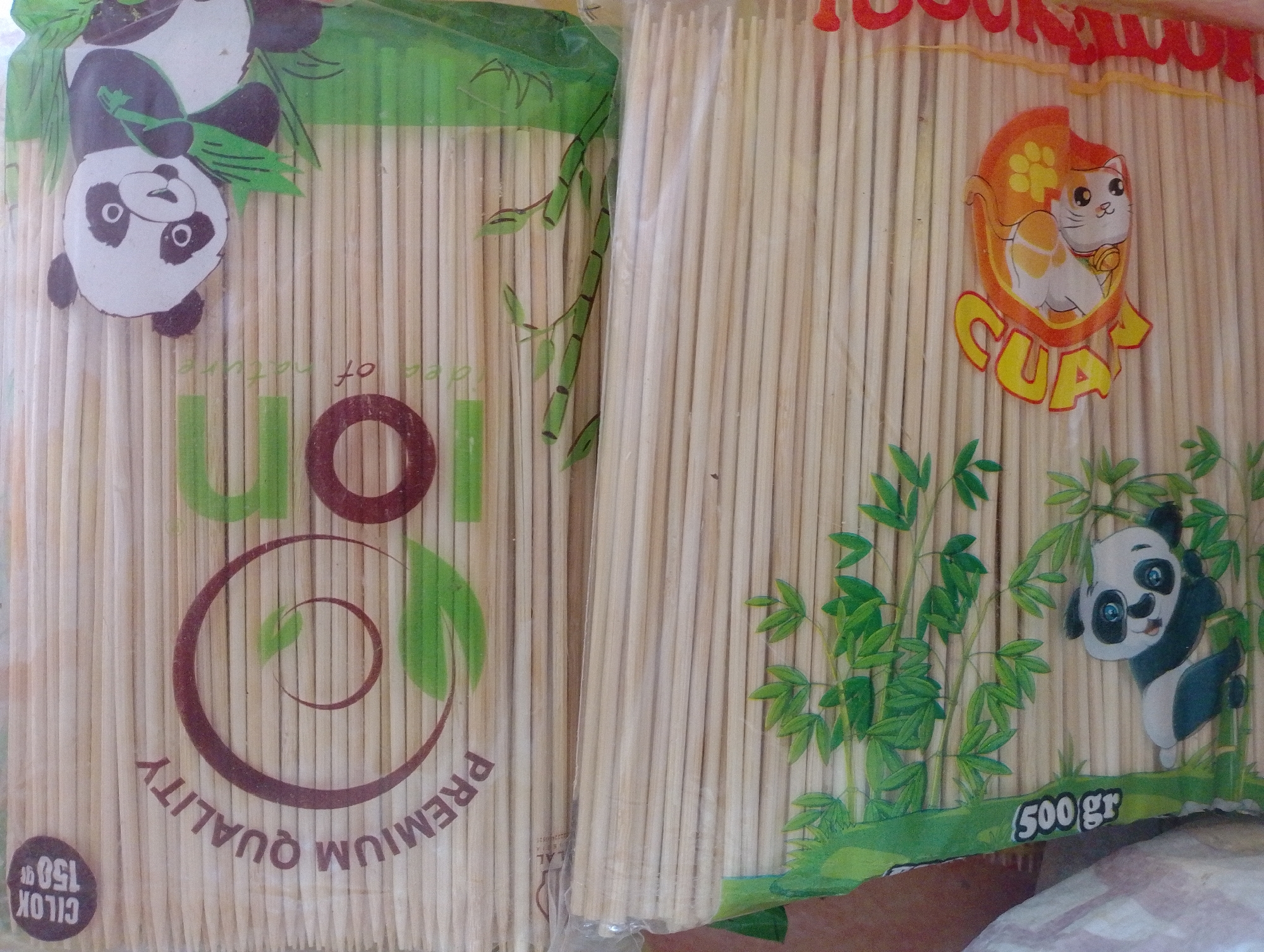This is a close-up, eye-level, medium shot of two plastic-wrapped packages of wooden skewers. The package on the left features a cartoon panda hanging upside down from bamboo stalks. Text around the panda includes "PREMIUM QUALITY" and "CILOK 150 gr". The package on the right displays a cartoon cat with orange and white fur, sitting in a round, orange graphic with a yellow paw print. Text below the cat reads "CUAN" in yellow lettering. Both packages are filled with numerous wooden skewers arranged vertically. The background is out of focus. The image was taken in Tungkaran Pangeran, Indonesia.