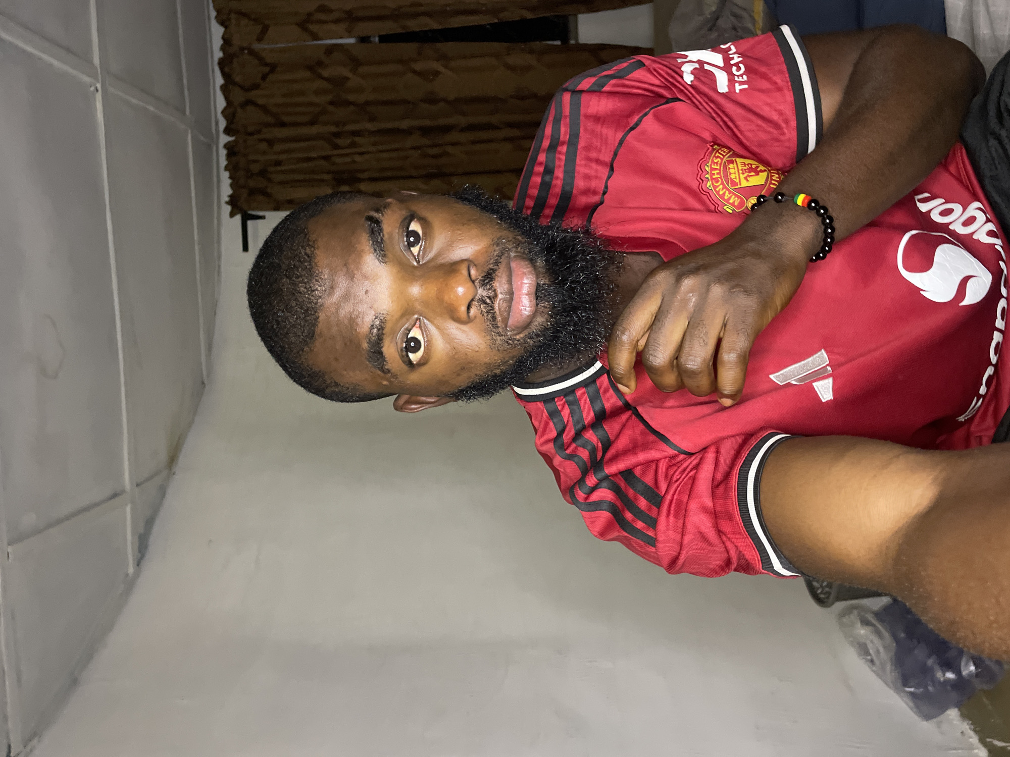 In a dimly lit room, a young Black man with a full beard and short hair is captured in a close-up, head-and-shoulders shot. He is wearing a red Manchester United jersey with black stripes on the shoulders and white trim on the collar and sleeves. His left hand rests on his chin, and he wears a black beaded bracelet with red, yellow, and green beads on his right wrist. The background is indistinct, featuring a textured wall and dark brown curtains. The ceiling appears to be a drop ceiling with visible tiles and some water staining. To the left, a plastic bag filled with blue material is partially visible, along with a dark cylindrical object that could be a trash bin. The overall ambiance suggests a casual indoor setting, possibly a bedroom, in Jos, Nigeria.