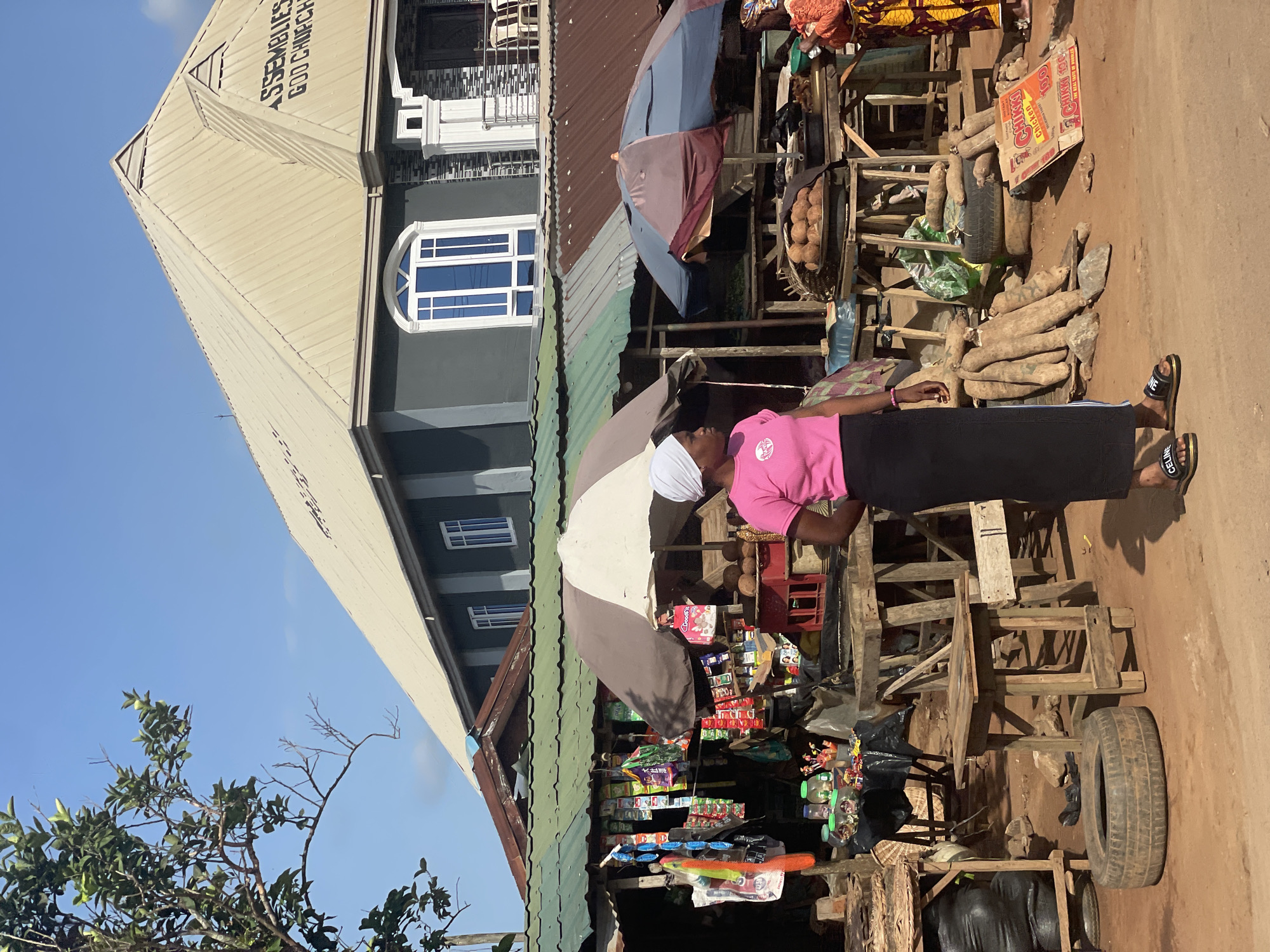 In Ipota, Nigeria, a woman wearing a pink shirt and black skirt stands at a roadside stall. The stall is covered by a striped umbrella and is stocked with various goods including snacks, packaged foods, and personal care items. Behind her, there are piles of yams and coconuts. The stall is constructed with wooden materials and incorporates old tires as part of its structure. In the background, there is a building identified as an Assemblies of God Church. The sky is clear and blue.